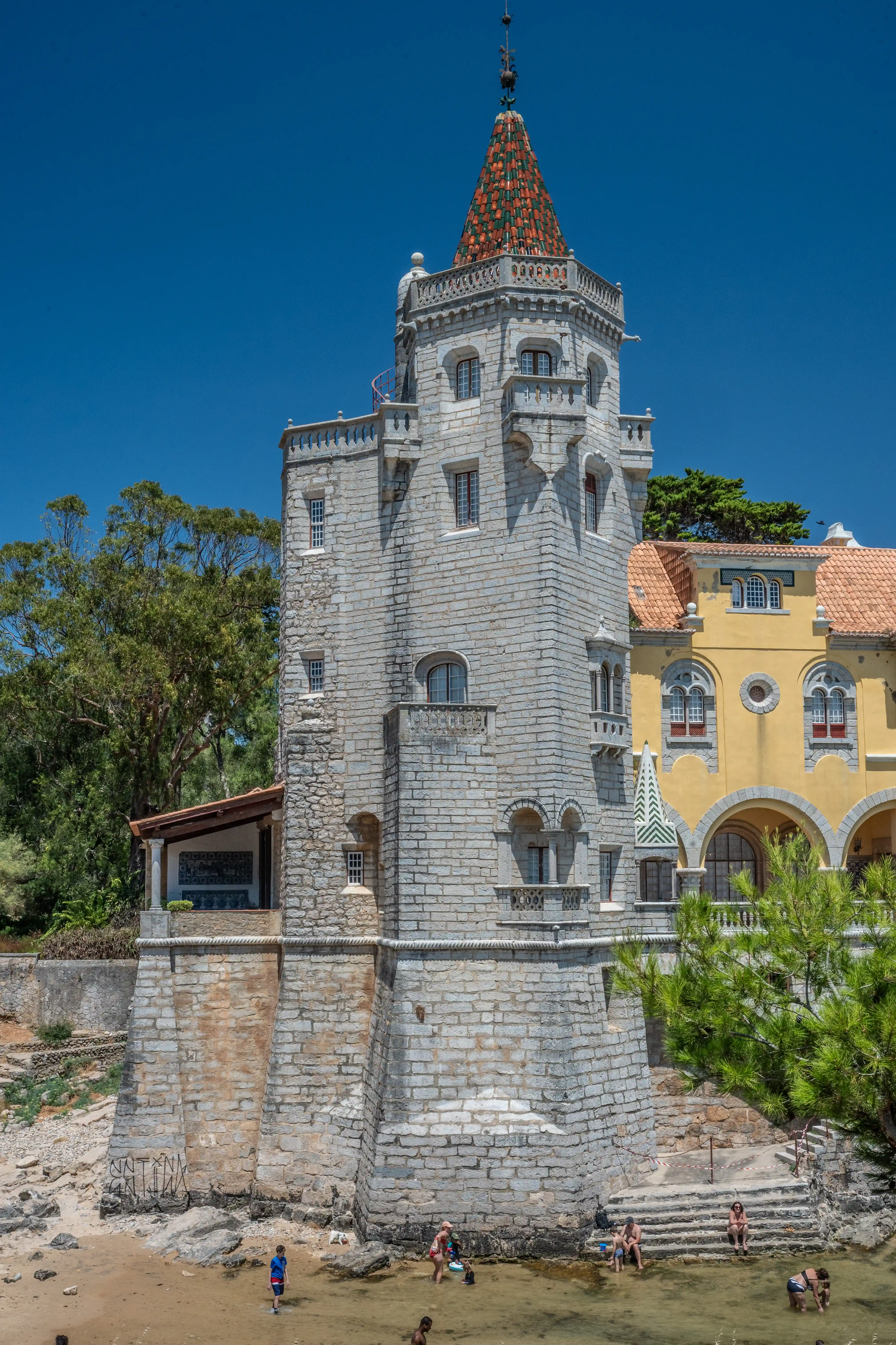A stone tower with a red and green tiled conical roof stands next to a yellow building with arched windows. People are visible wading and sitting near the water's edge on a sandy and rocky shoreline.