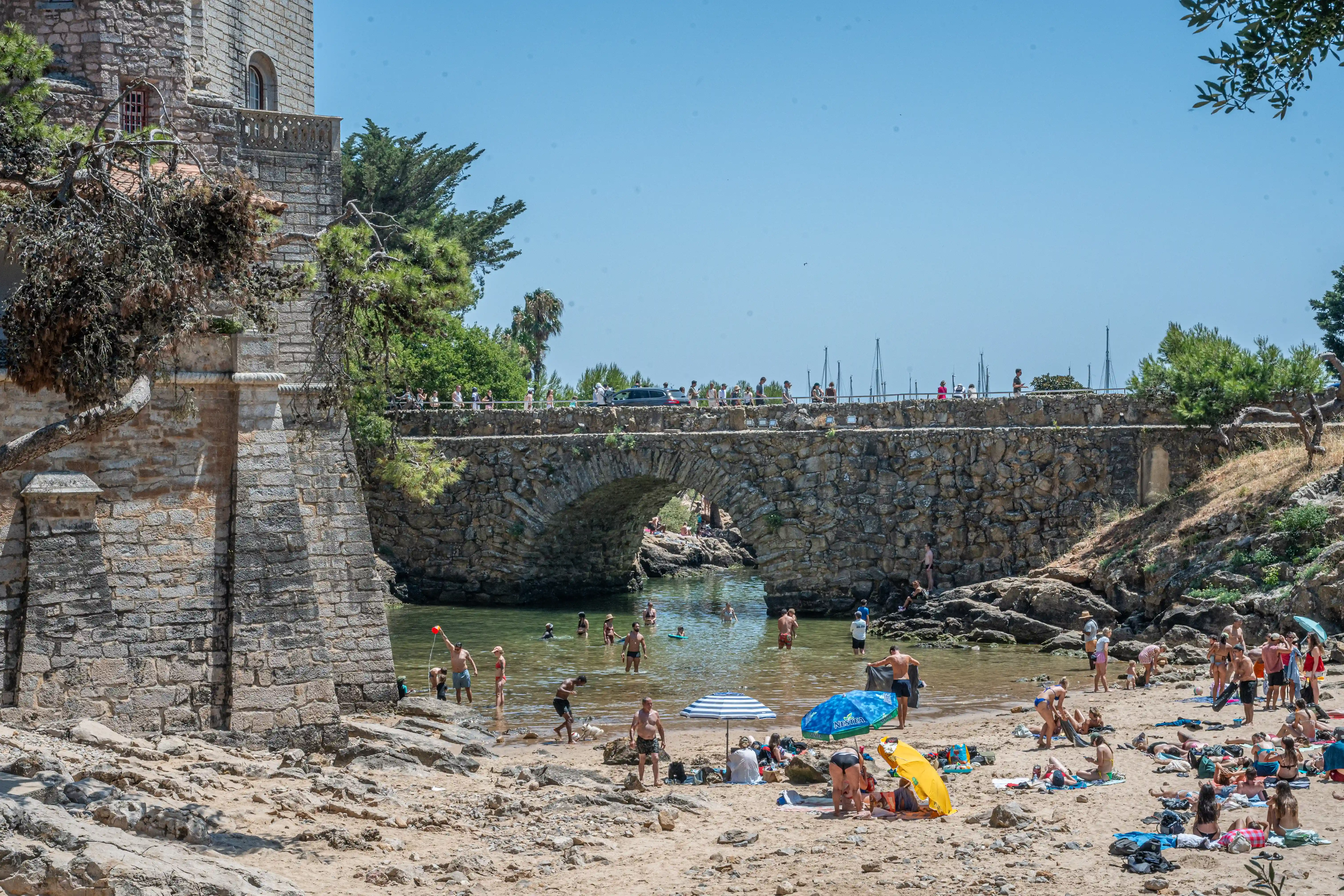 A crowded beach scene with people sunbathing on the sand and swimming in the sea, with a large, old stone bridge arching over the water in the background.