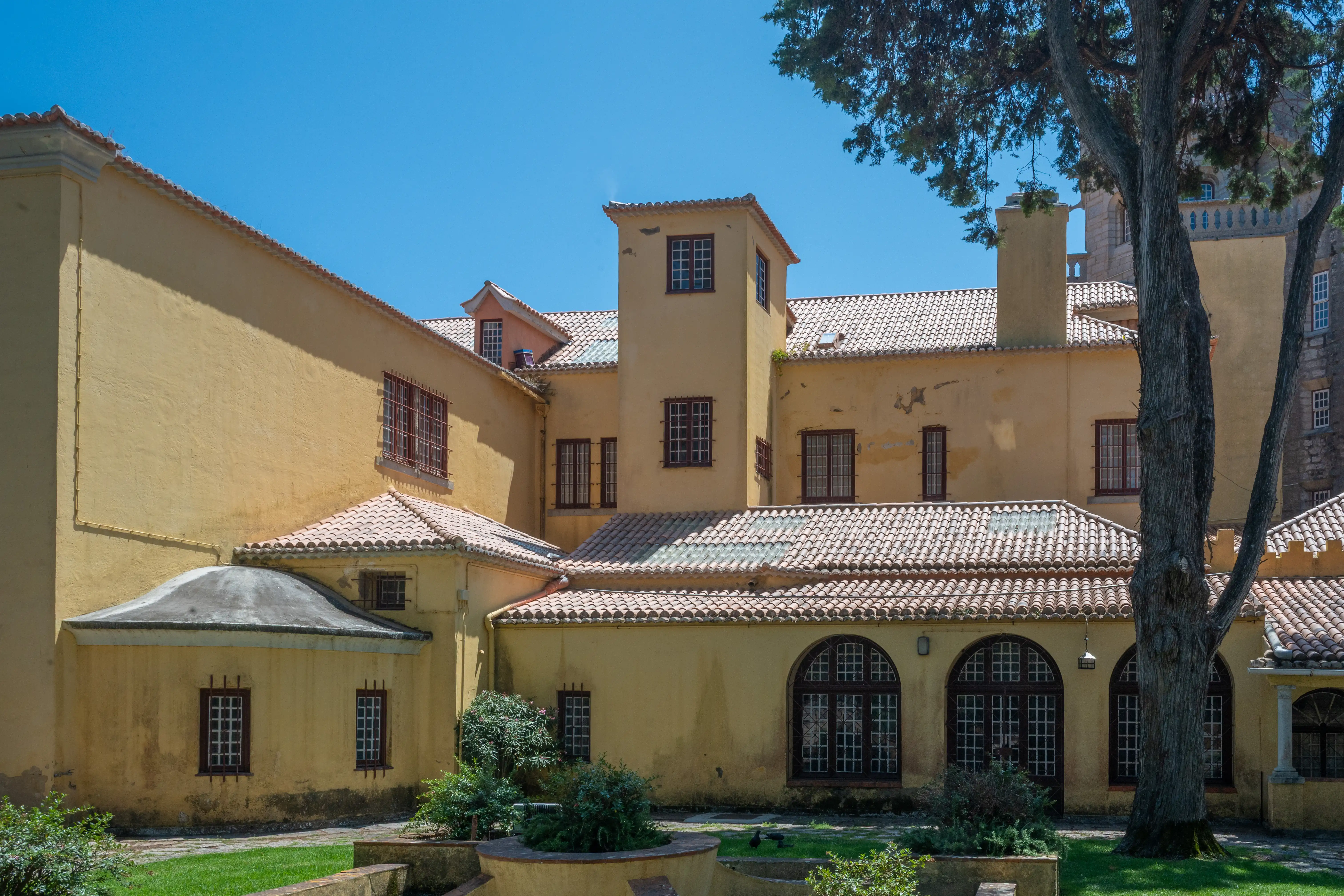 An exterior shot of a pale yellow building with many windows and a reddish-brown tiled roof, partially shaded by a large tree on the right. The building has a central tower and several wings. A green garden area is in the foreground.