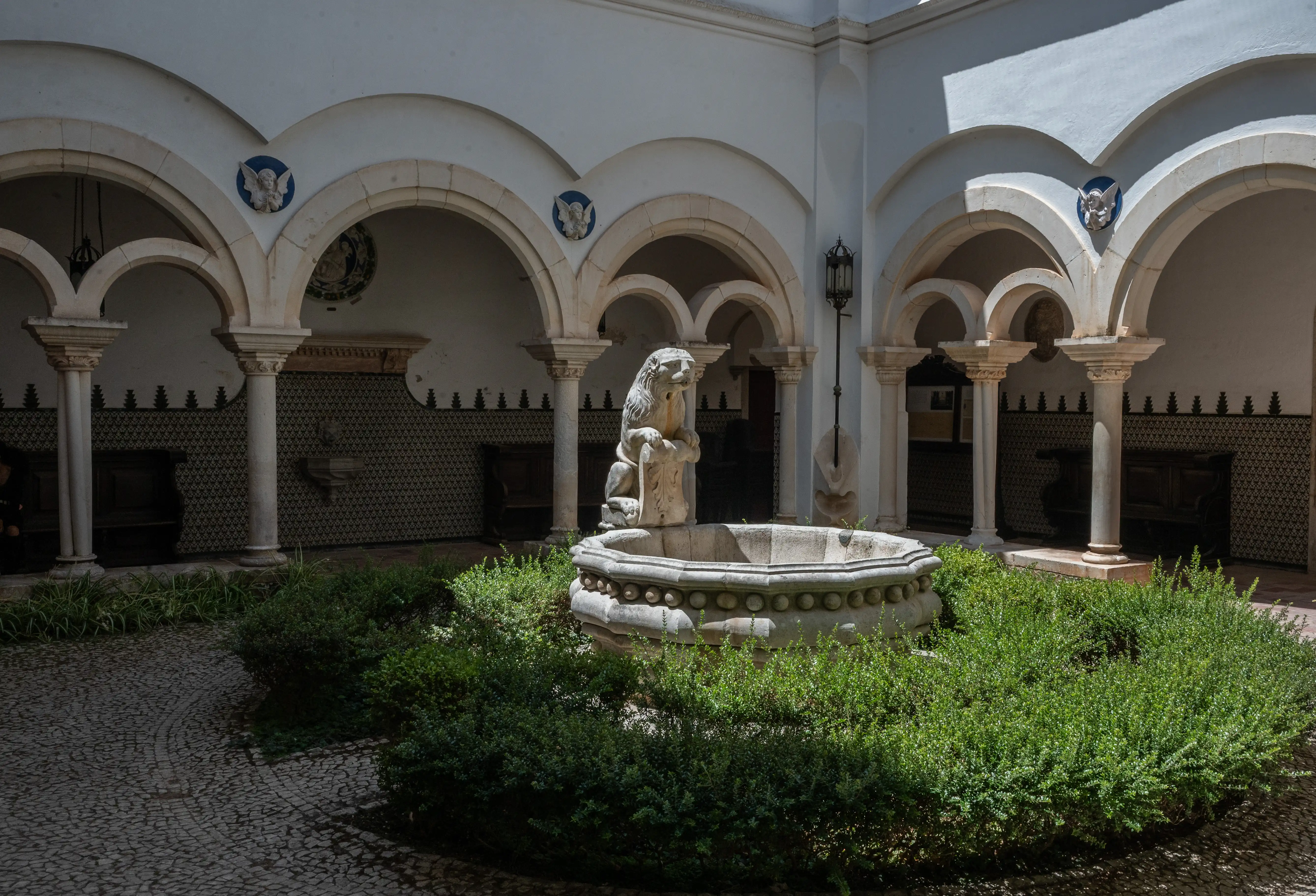 Stone lion statue on a fountain pedestal in a sheltered courtyard with white arches, columns and landscaped greenery.