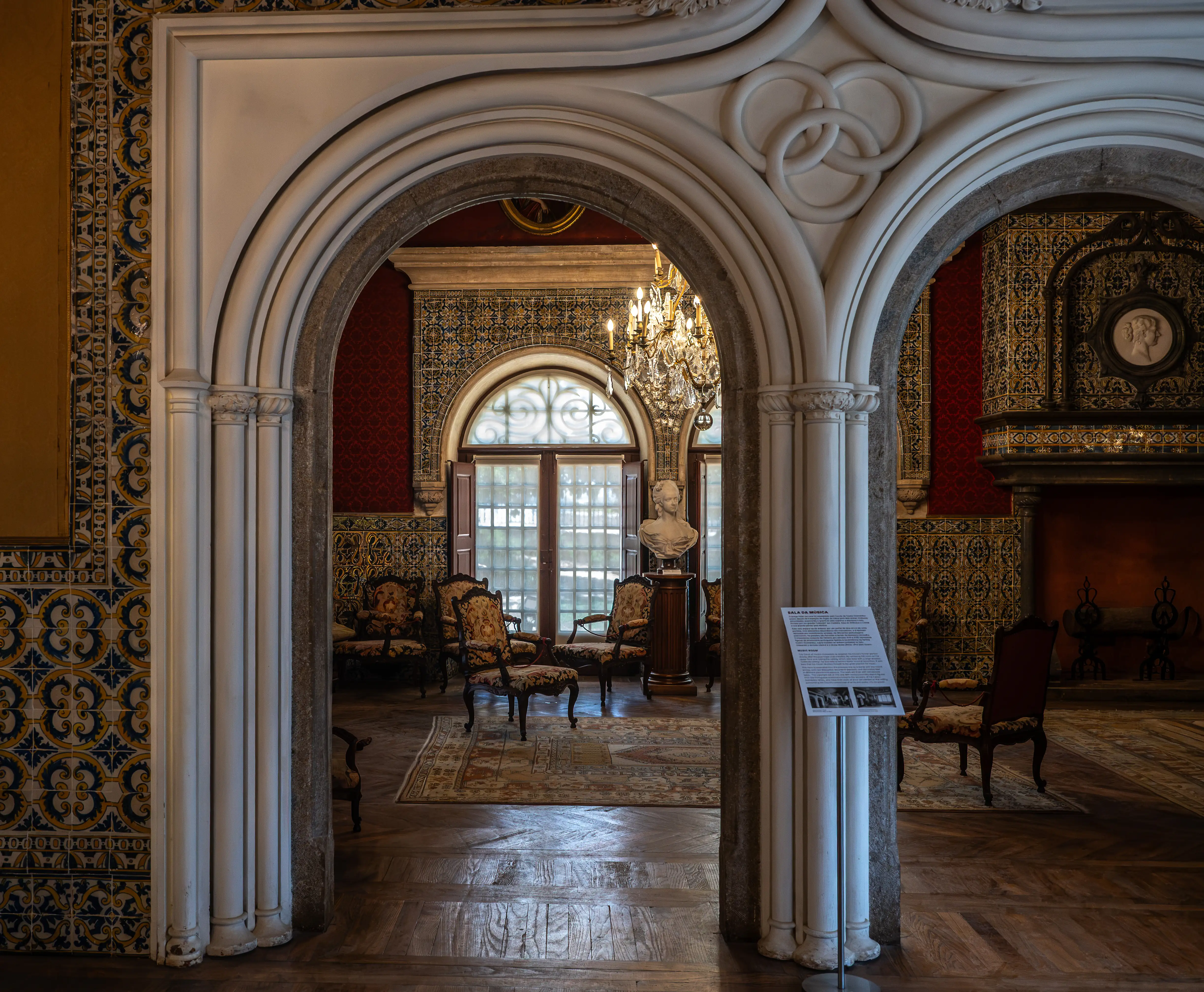 Interior view of a historic building, looking through a stone archway. Beyond the arch, a salon is visible with ornate furniture, patterned walls, a grand chandelier, and a large arched window. A person is seen standing to the right.