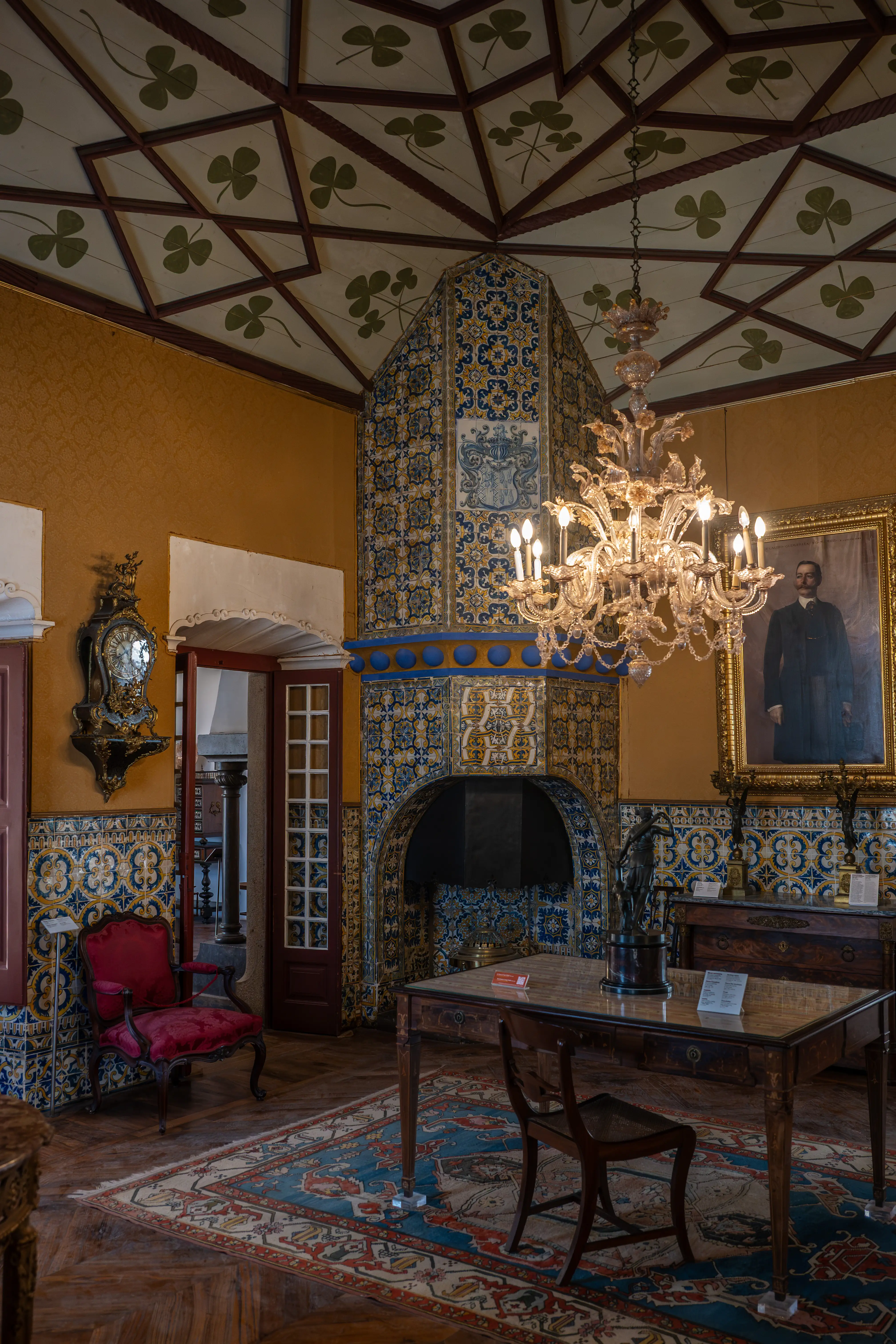 Interior of a historic room with walls and a fireplace covered in intricate blue and yellow azulejo tiles. A large crystal chandelier hangs from a decorated ceiling with a geometric pattern and green clover designs. A framed portrait of a man in formal attire hangs on the wall, alongside a decorative clock. In the foreground, a wooden table and chairs sit on a patterned rug, with a red velvet armchair to the left.