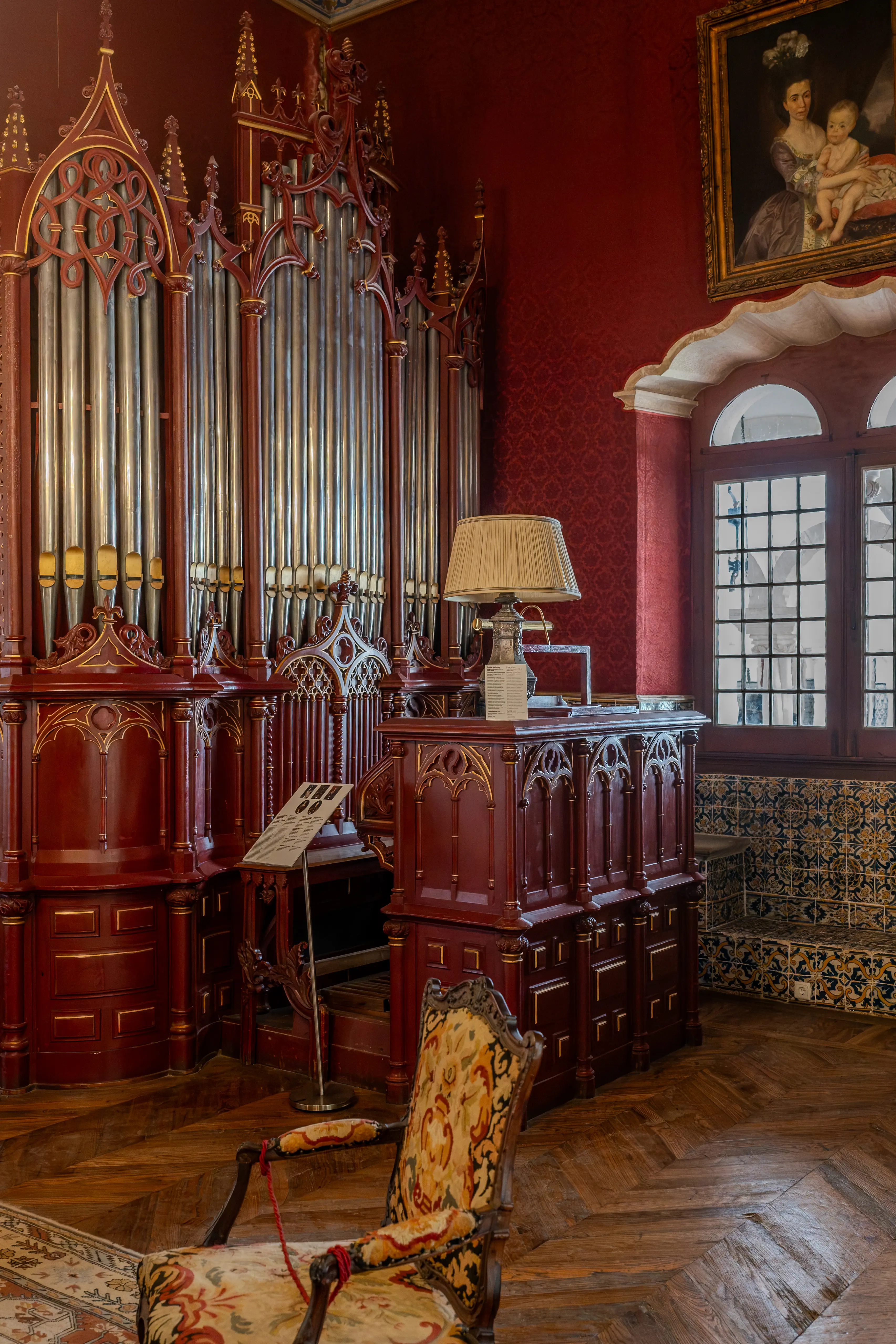 An ornate pipe organ with metallic pipes and dark red wooden casing stands in a richly decorated room. In the foreground, a vintage armchair with a colourful floral tapestry upholstery sits on a patterned rug. To the right, a pair of arched windows with leaded panes are visible, and a framed portrait hangs on the wall above them.