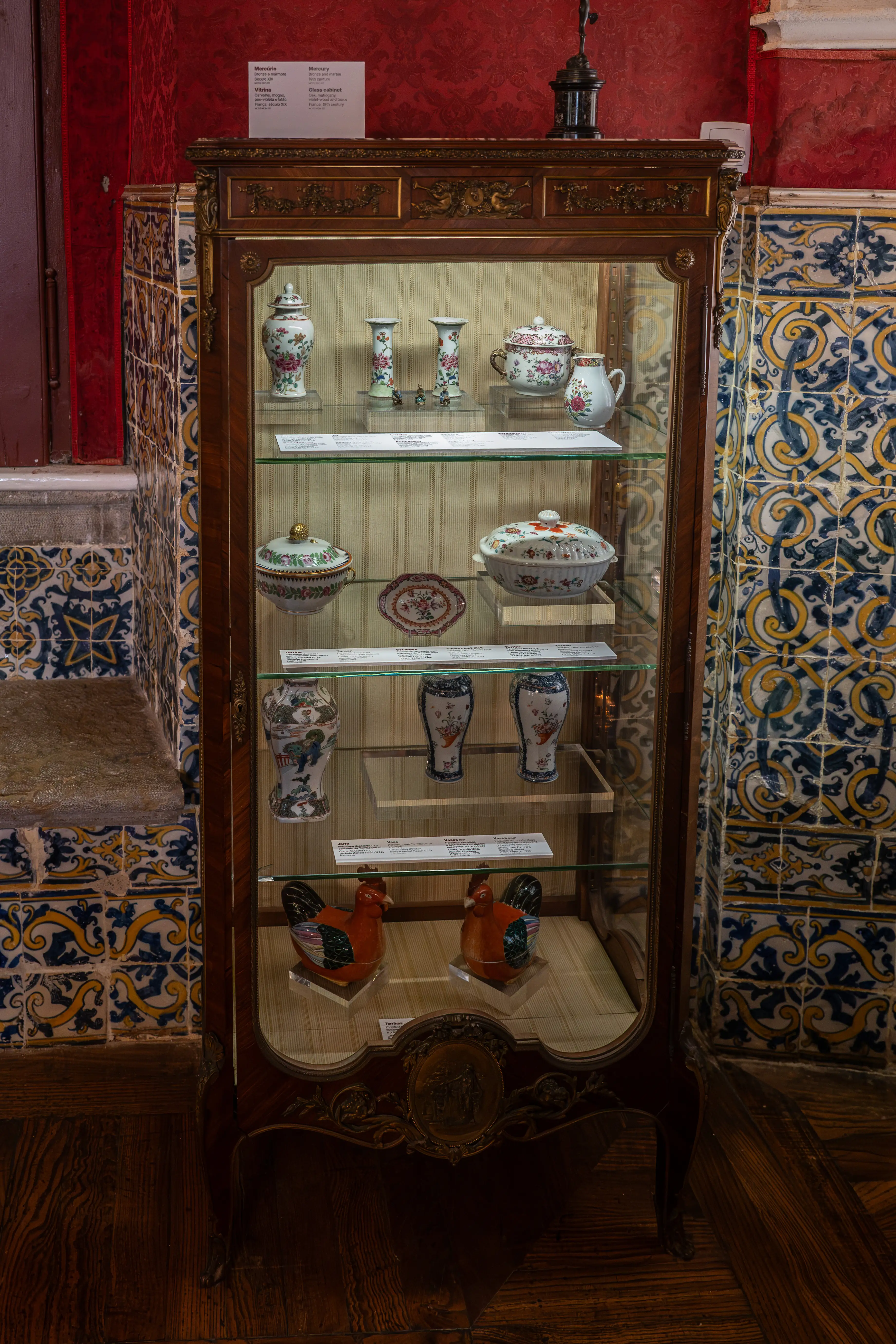 A museum display cabinet filled with various porcelain and ceramic pieces, including vases, a teapot, and decorative rooster figurines. The cabinet is surrounded by tiled walls.