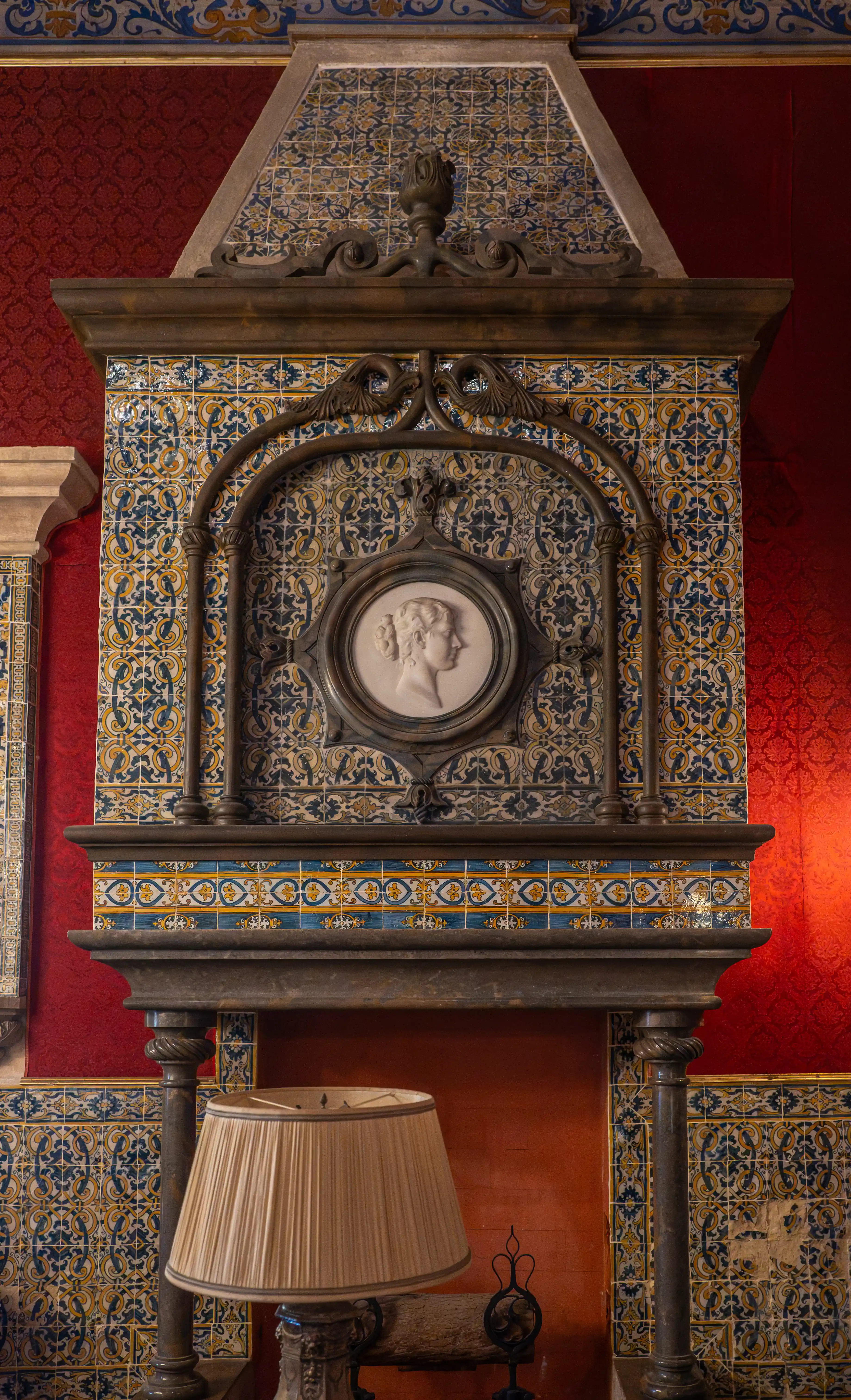 A large, ornate fireplace with colourful patterned tiles and a classical medallion above, set against a red patterned wall with a crystal chandelier.