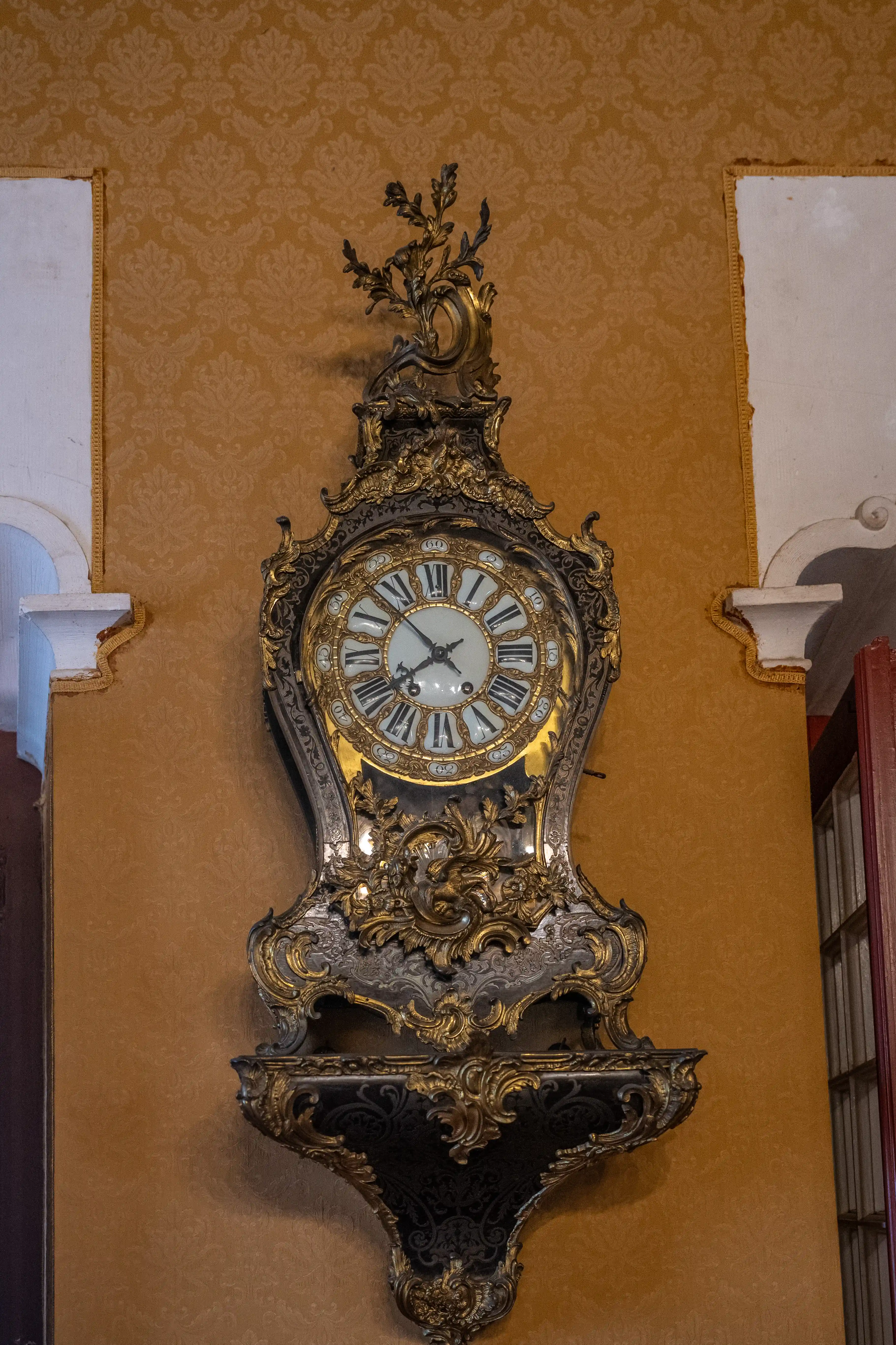 An antique, gilded wall clock with Roman numerals is displayed on a patterned ochre wall, featuring ornate metalwork and a decorative shelf.