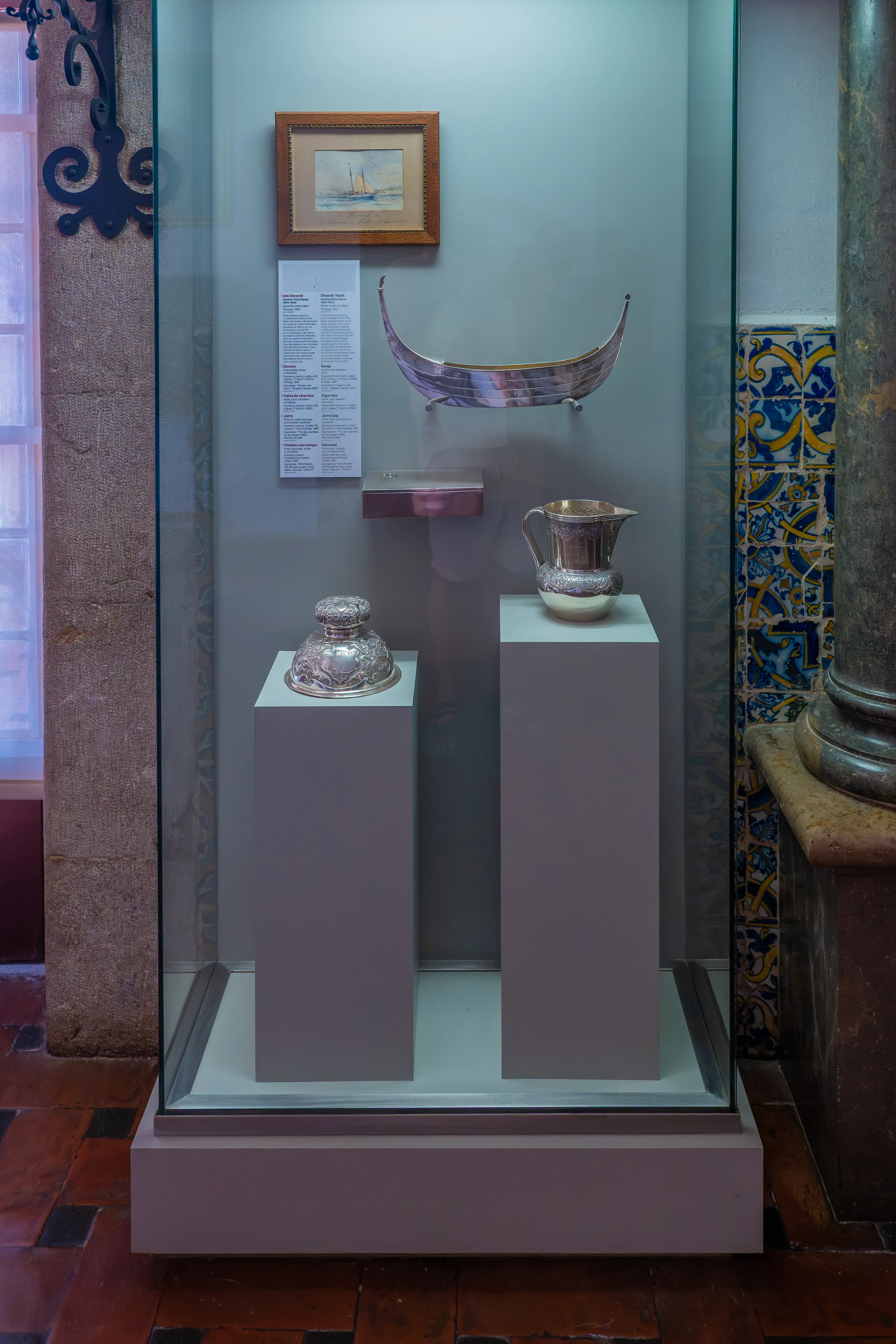 A museum display case with several artefacts, including a small framed painting of a boat, a silver decorative boat-shaped object, a silver inkwell with intricate detailing, and a silver jug, all arranged on plinths.