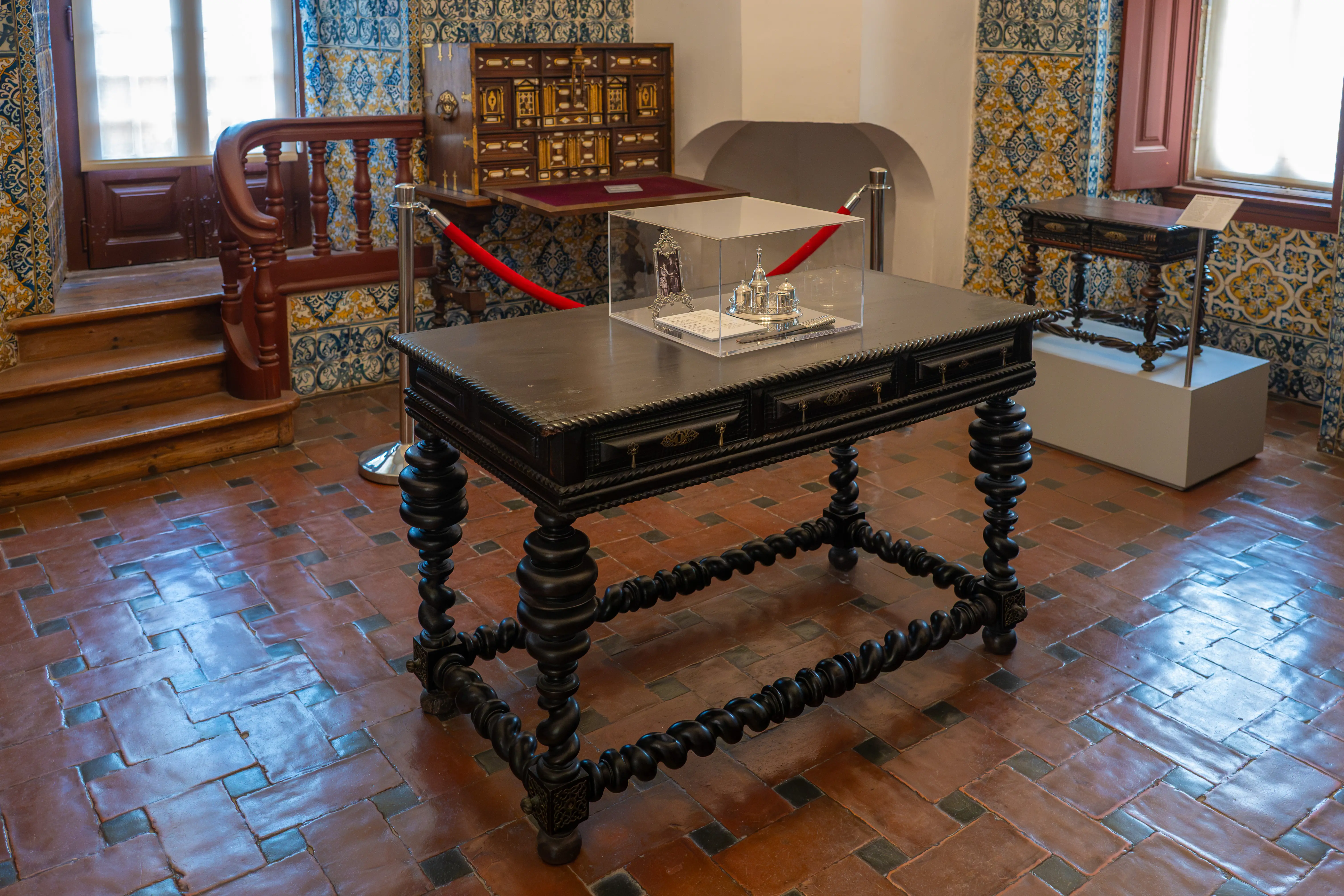 A dark wooden table with intricately carved, twisted legs displayed in a room with decorative blue-and-white tiles. A glass display case with small ornate objects is visible in the background