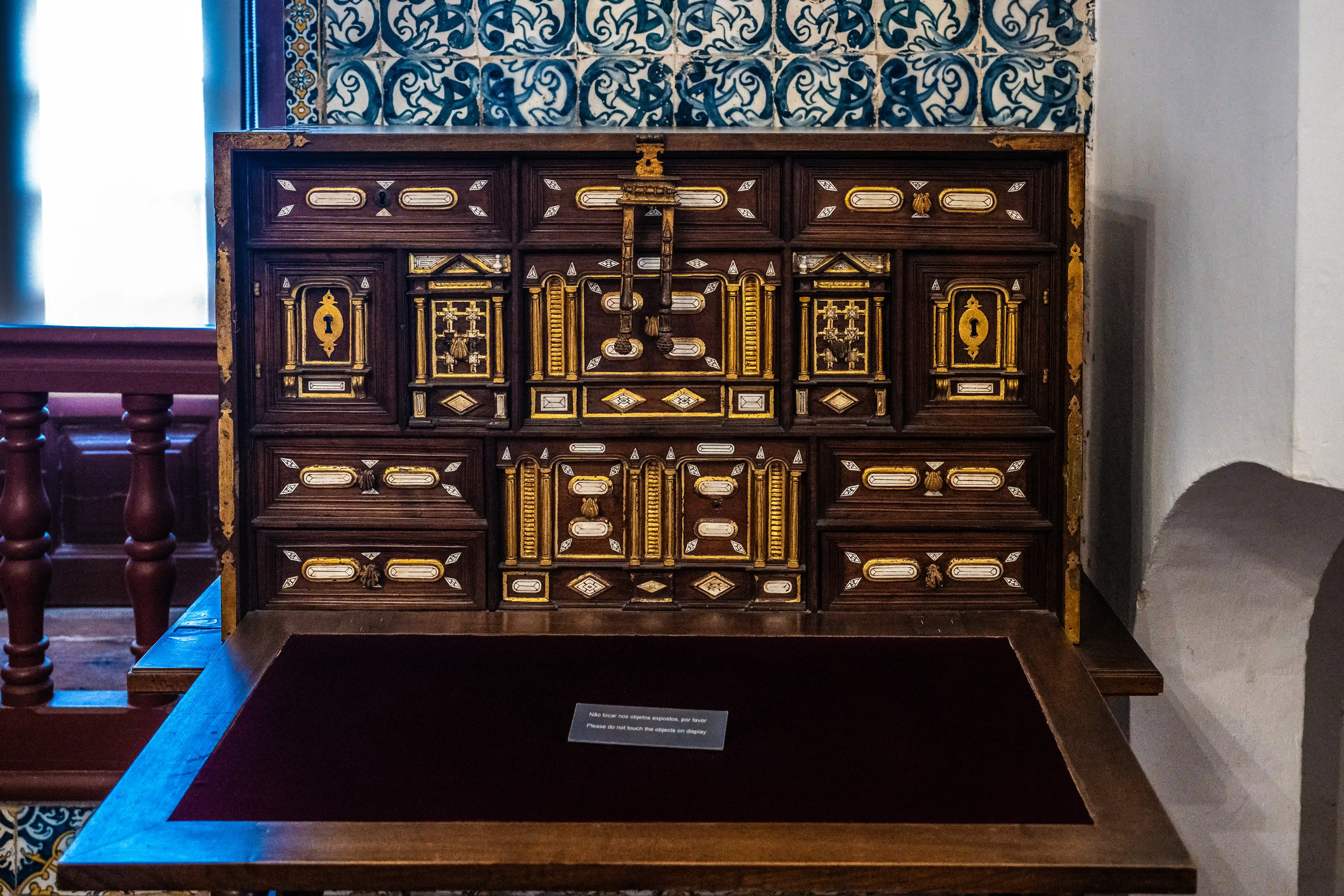 An antique wooden bureau desk with a dark brown finish, adorned with elaborate gold and white decorative inlays and small drawer fronts. The desk is open, revealing a burgundy velvet writing surface. Behind the desk, a wall is decorated with blue and white patterned tiles.