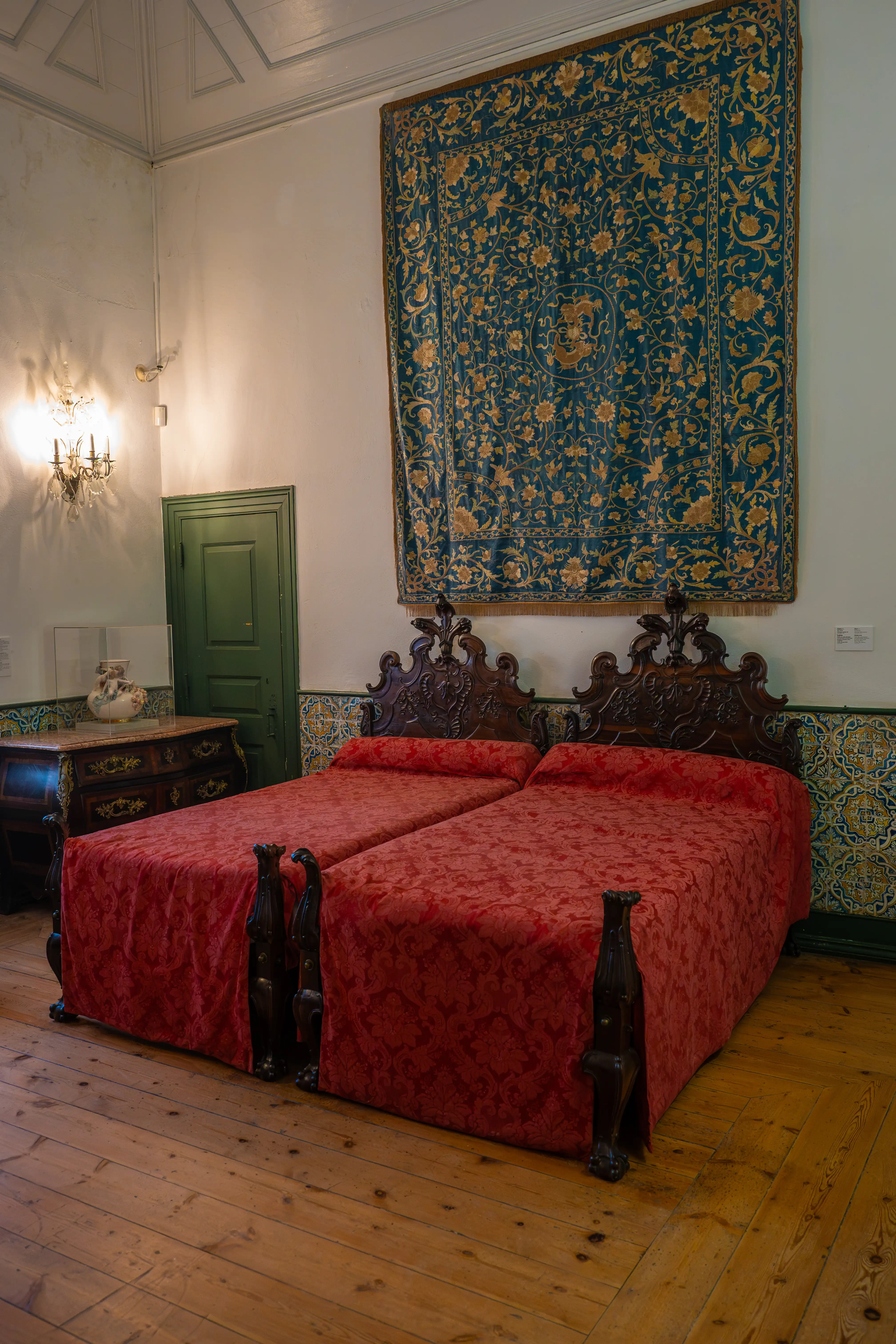 A bedroom with two twin beds covered in red fabric, dark wooden headboards, and a large tapestry on the wall.