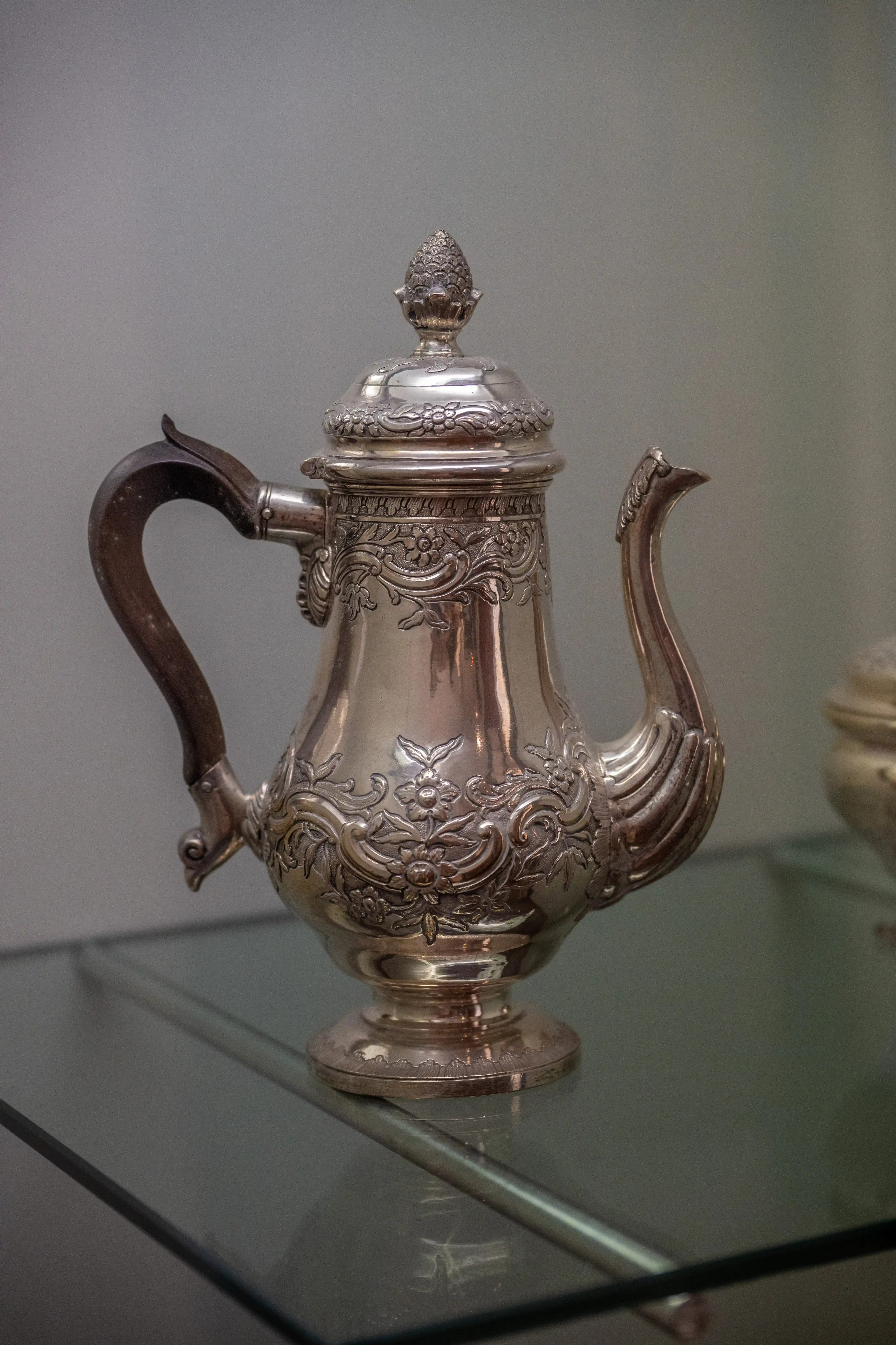 A close-up of a silver teapot with intricate floral and scrollwork engravings. The teapot has a curved handle, a finial on the lid, and a detailed spout, with a polished surface that reflects light