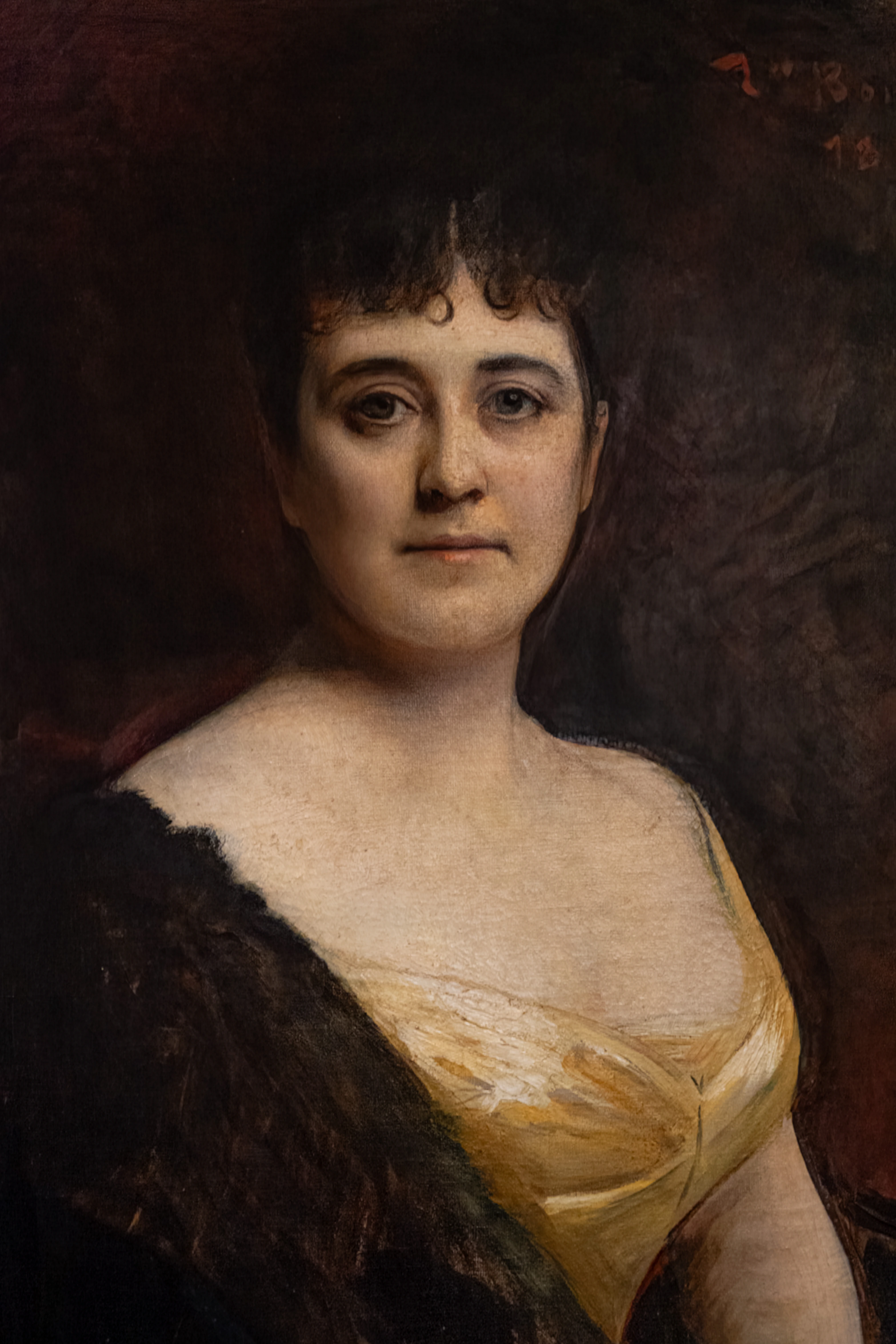 Oil painting portrait of a woman looking directly at the viewer. She has dark hair with bangs and curls on her forehead, fair skin, and is wearing a yellow dress with a low neckline and a black shawl.