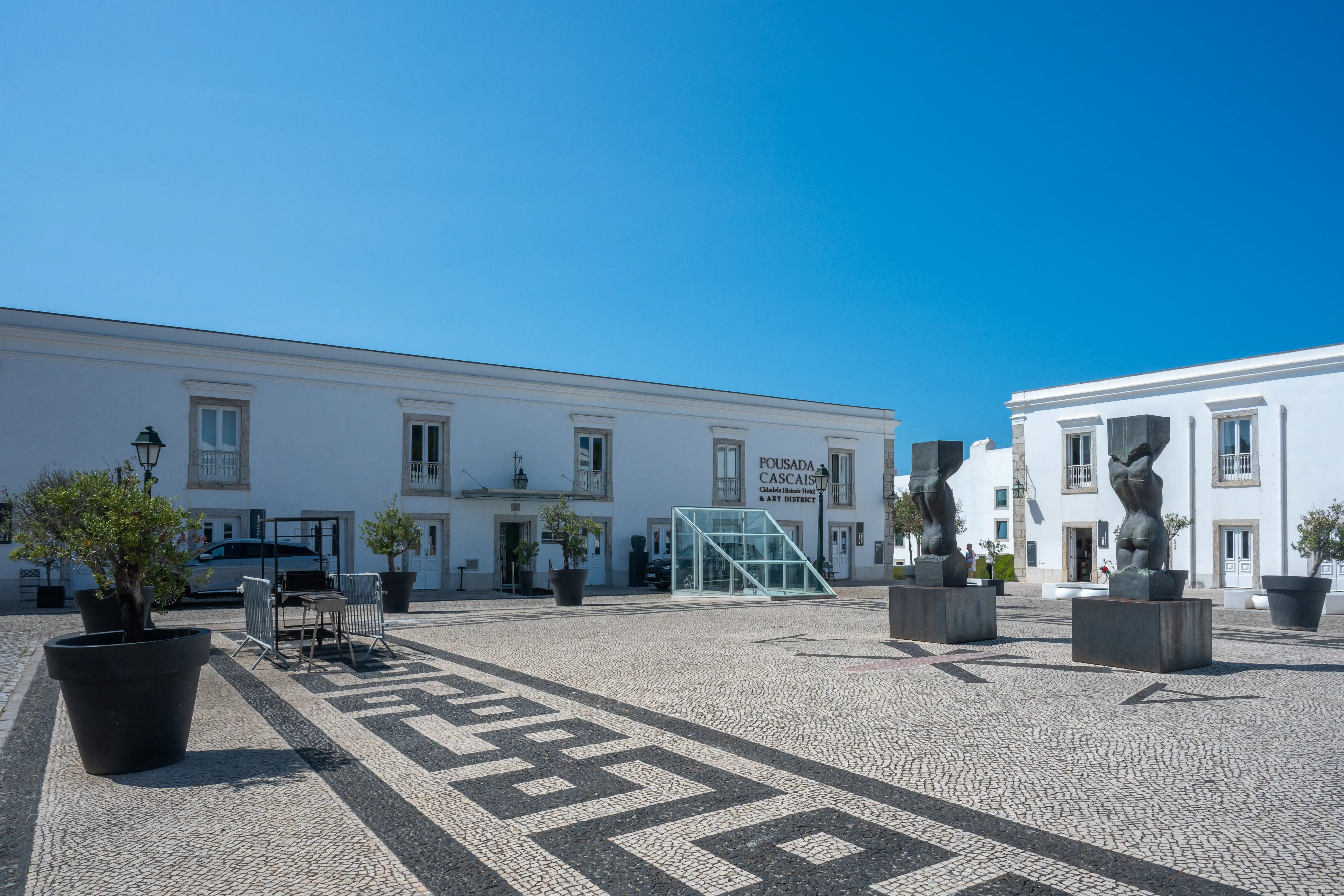 Two dark, abstract human-like sculptures with blocky heads are positioned in a public square. The square is paved with a black and white mosaic pattern. White buildings, including one with a sign for 