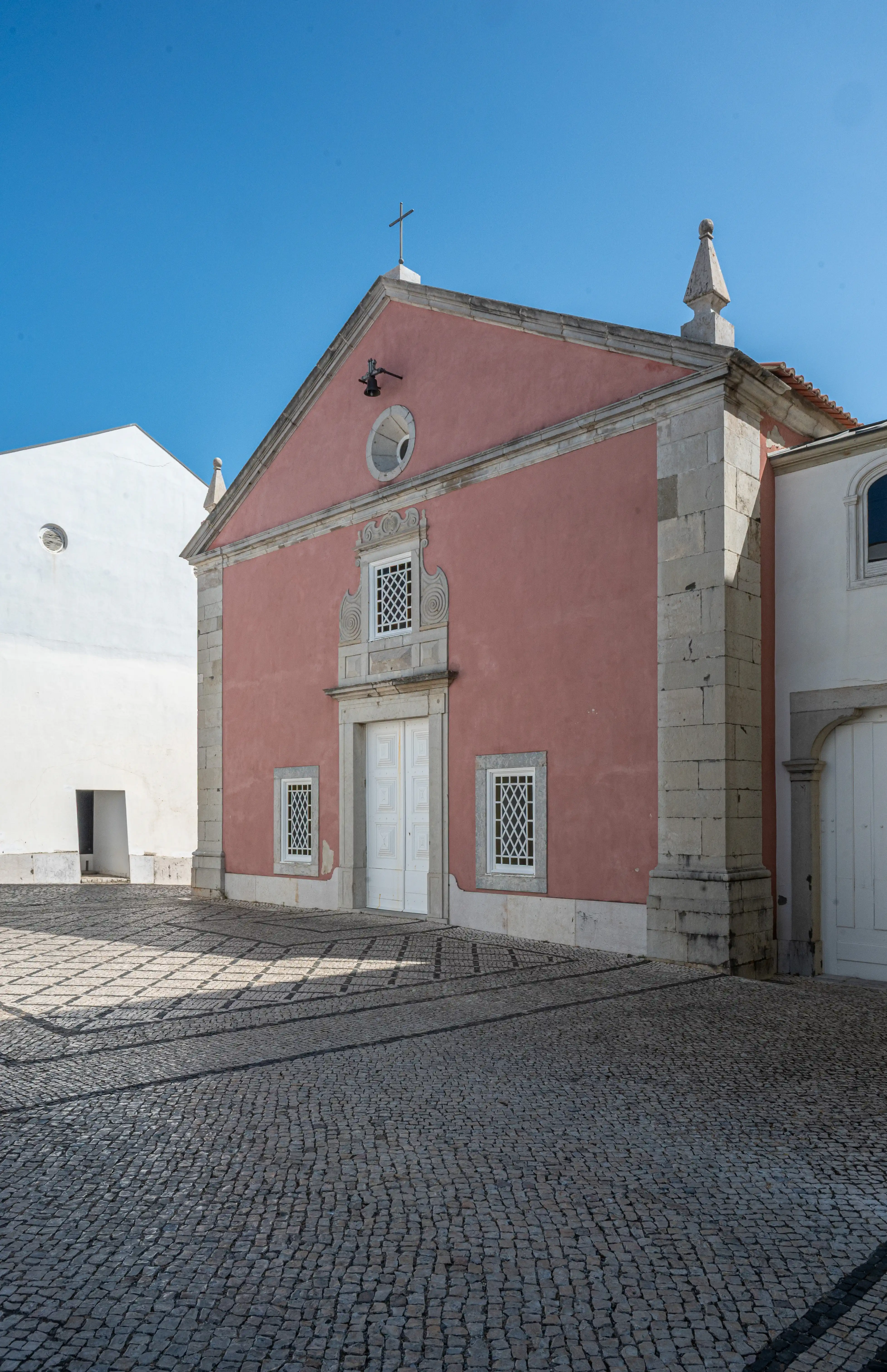 An upward view of a building with a pink façade and white-trimmed windows featuring lattice designs. A textured stone wall is visible to the left, and the sky is clear and blue.