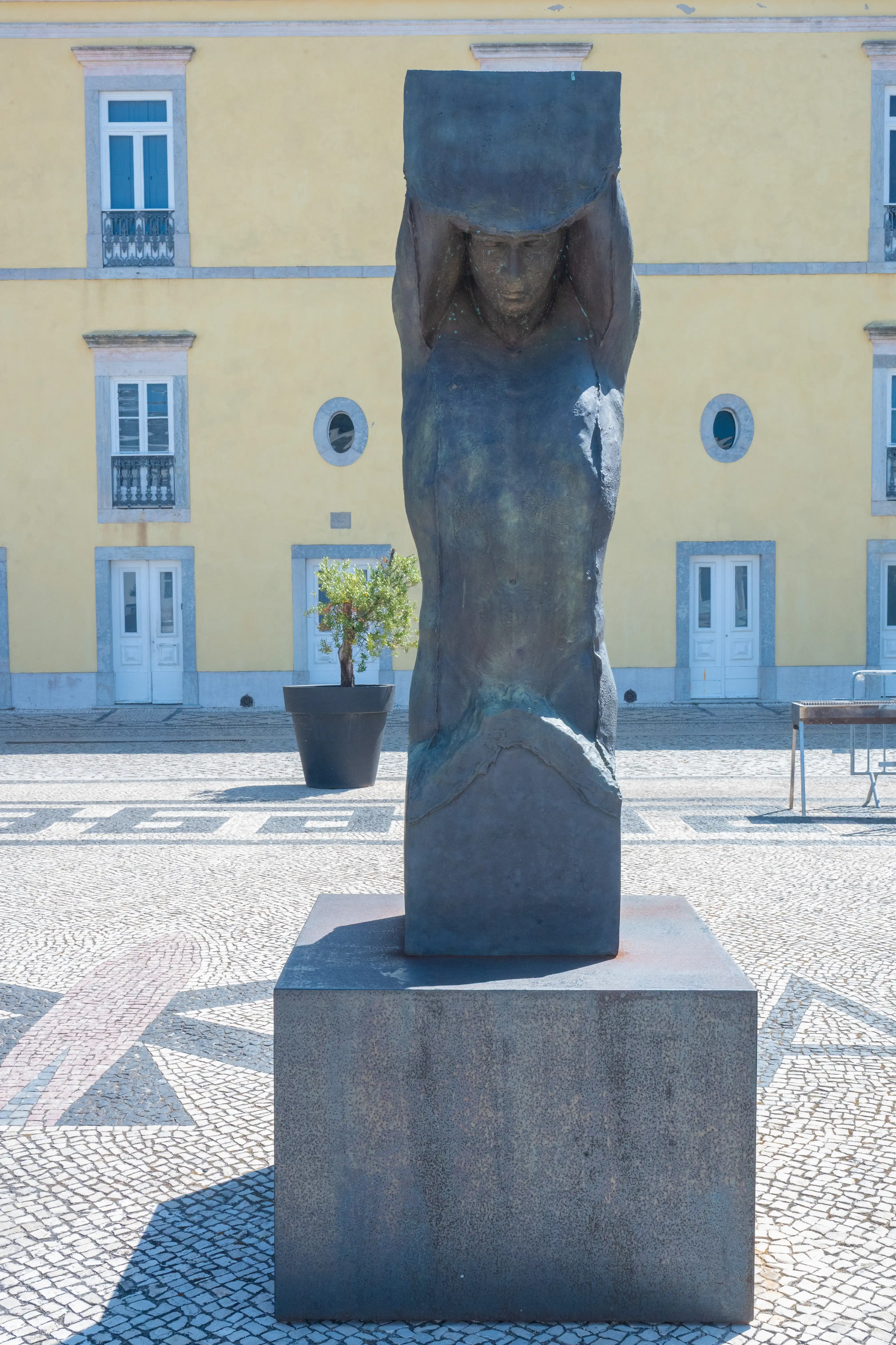 An aerial view of a bronze statue of a seated figure on a large stone pedestal in an urban square. The statue has a greenish patina, and the square features cobblestone patterns, a tree in a planter, and a pale yellow building with white-trimmed windows