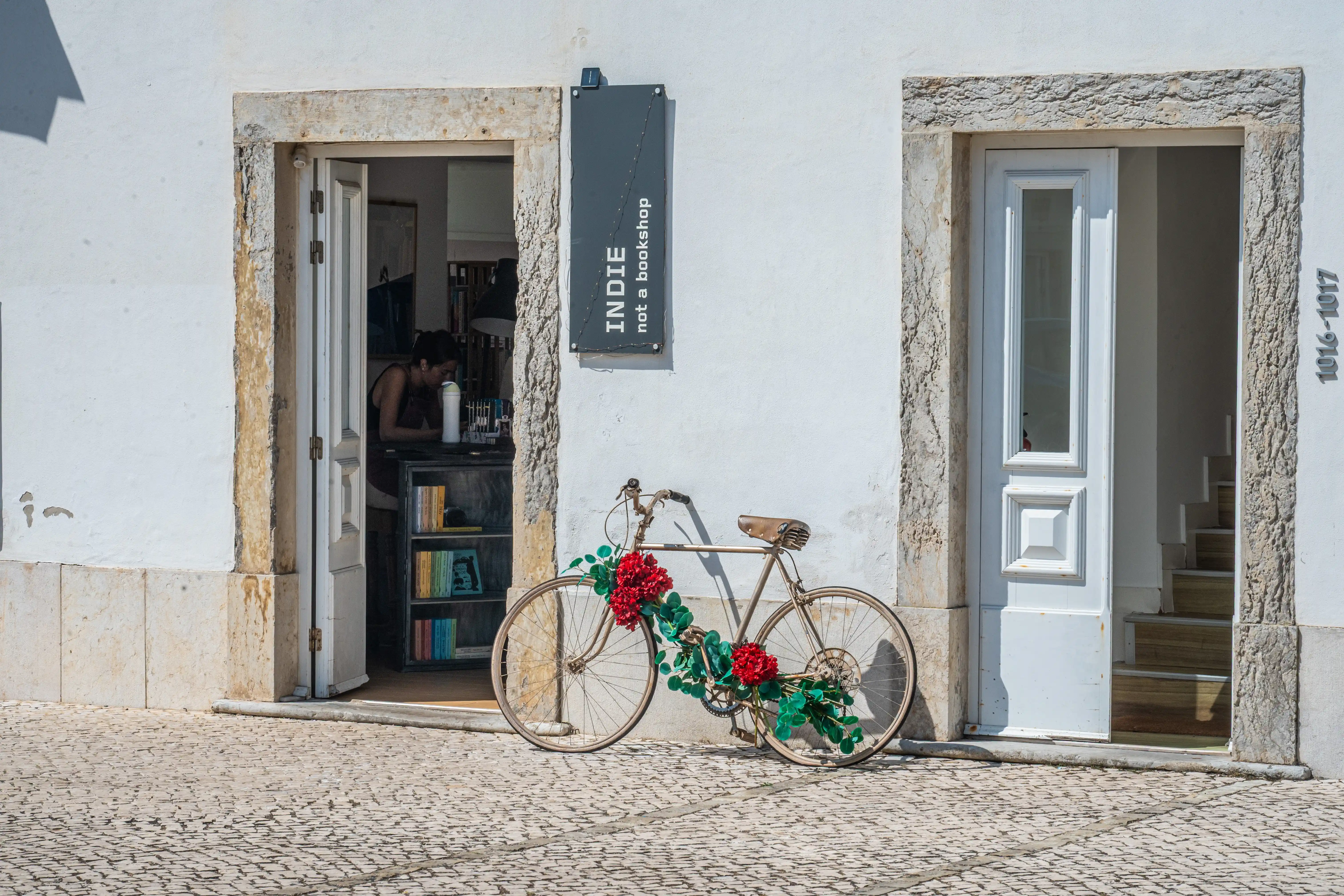 A vintage bicycle decorated with red flowers and green foliage stands in front of a white building with two doorways. One doorway is open, showing bookshelves inside a bookshop named 'Indie not a bookshop'
                         >
                         <figcaption>
                            <meta itemprop=