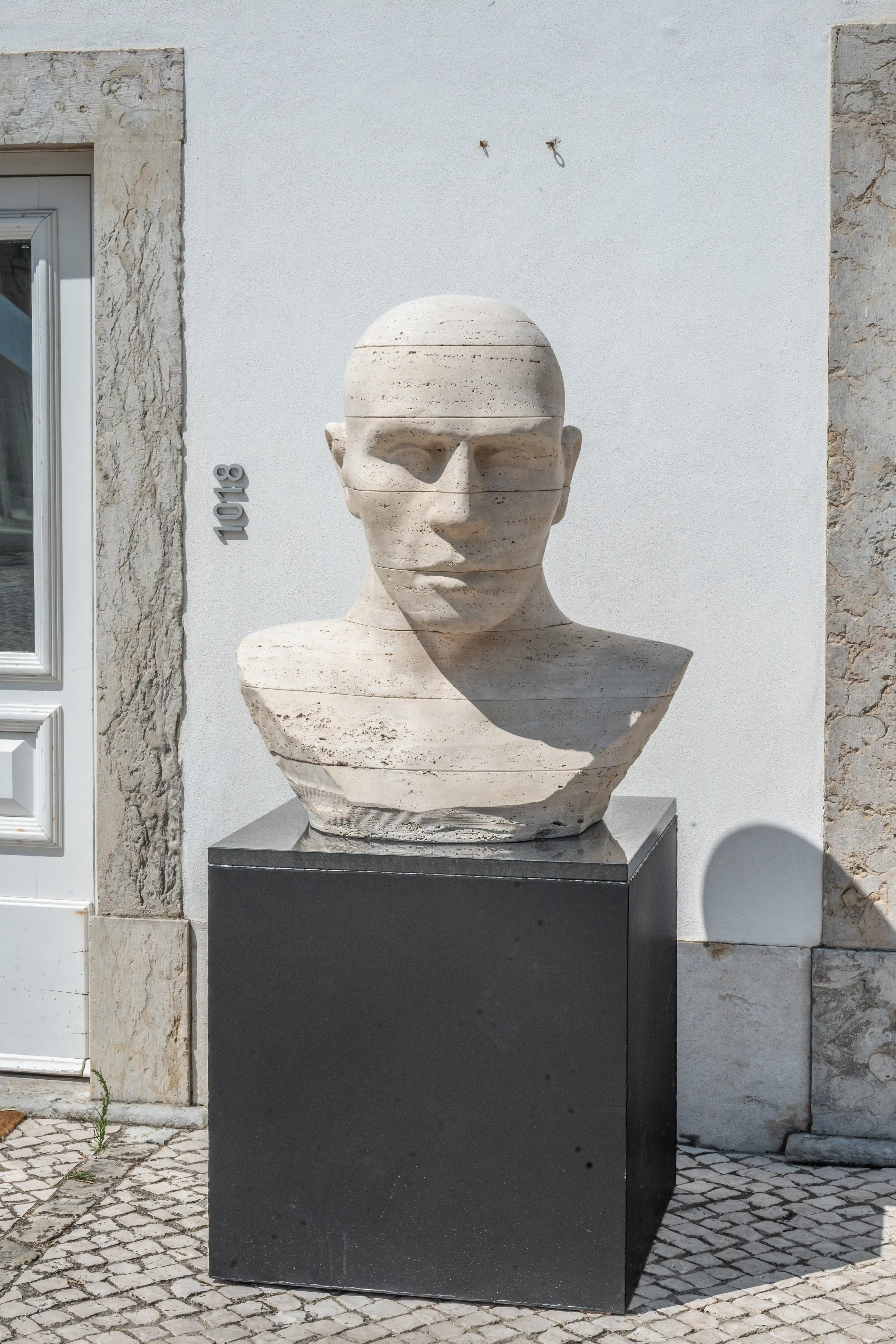 A light-coloured, horizontally layered stone bust of a person stands on a black cube pedestal. The sculpture is positioned outdoors in front of a white wall with textured stone details and a white door. The number '1018' is visible on the wall.