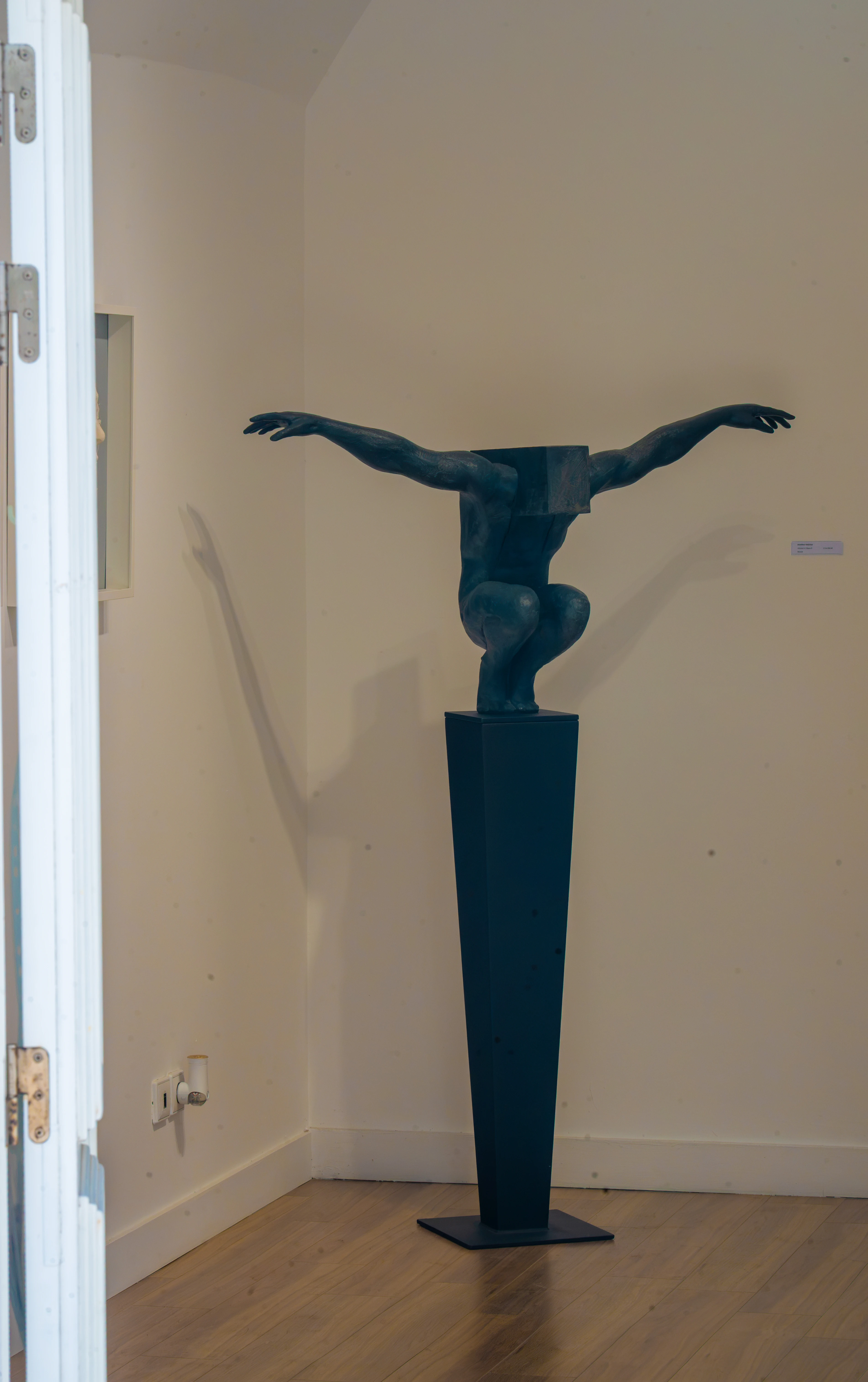 A dark bronze abstract sculpture of a person crouching with their arms spread wide, their head replaced by a cubical form. The sculpture stands on a tall, dark, angular plinth in an art gallery.