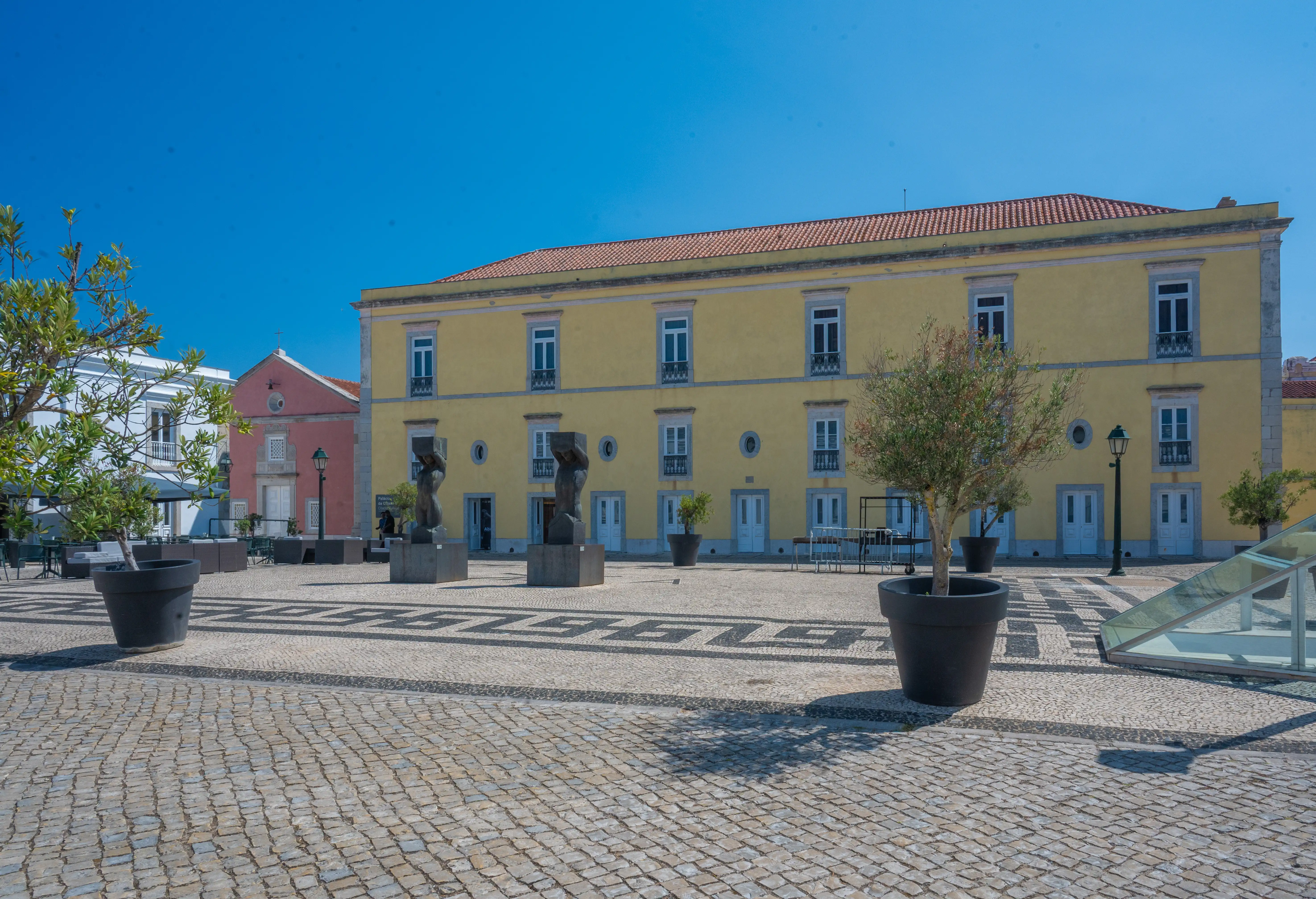 A plaza paved with cobblestones and black-and-white decorative tiles. A large pale yellow building with white-trimmed windows and a red-tiled roof stands in the background. Modern sculptures, potted trees, and outdoor seating areas are visible, and the sky is clear and blue.