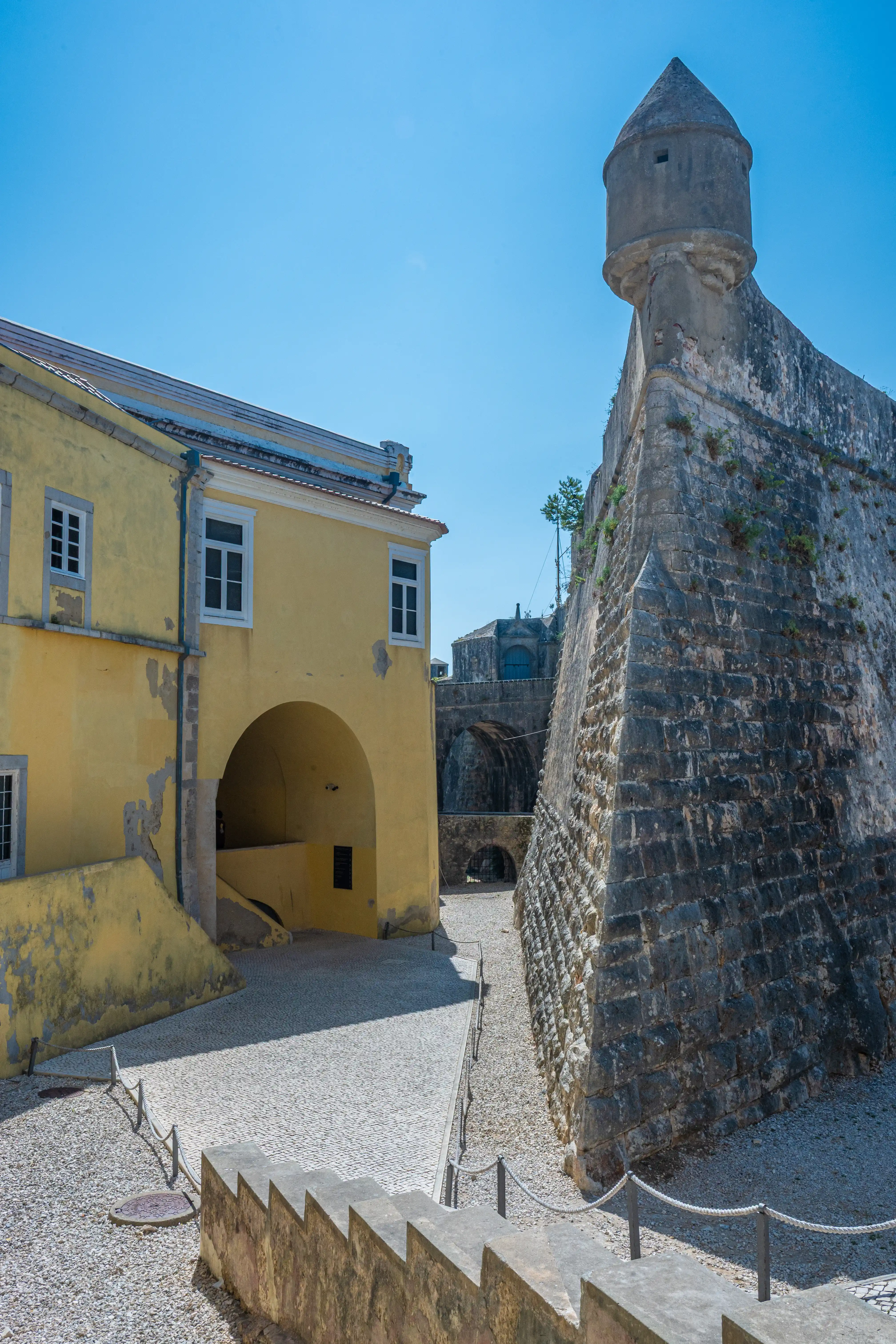 A historic stone fortification with a conical turret is seen on the right, with a pale yellow building featuring white-framed windows and an arched entrance on the left. The ground is a mix of gravel and cobblestone, with rope barriers and stone steps in the foreground.