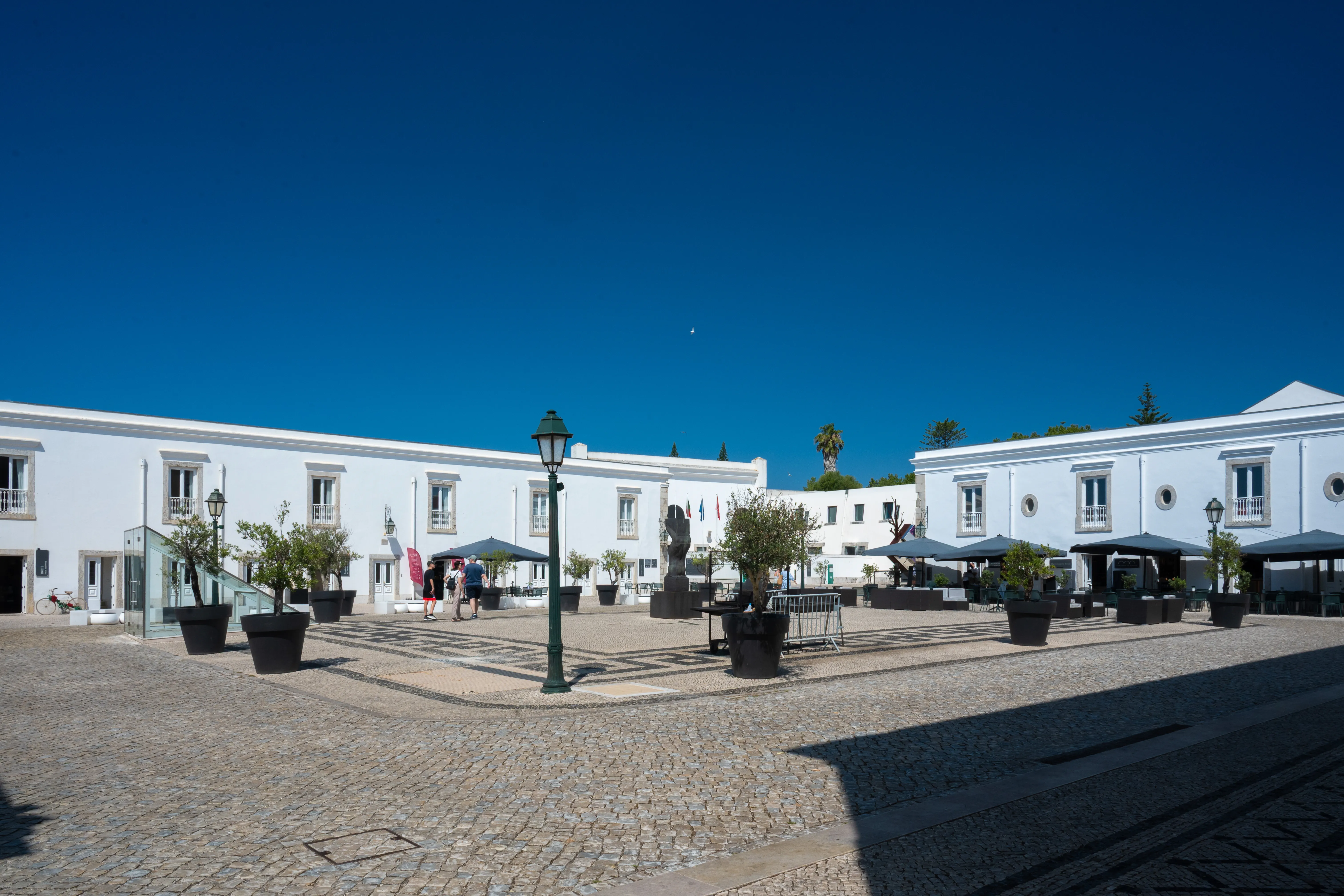 A spacious courtyard paved with cobblestones and black-and-white decorative tiles. White buildings with dark doors and windows surround the plaza. Potted trees and outdoor seating areas with umbrellas are visible, and a few people are walking around under a clear blue sky