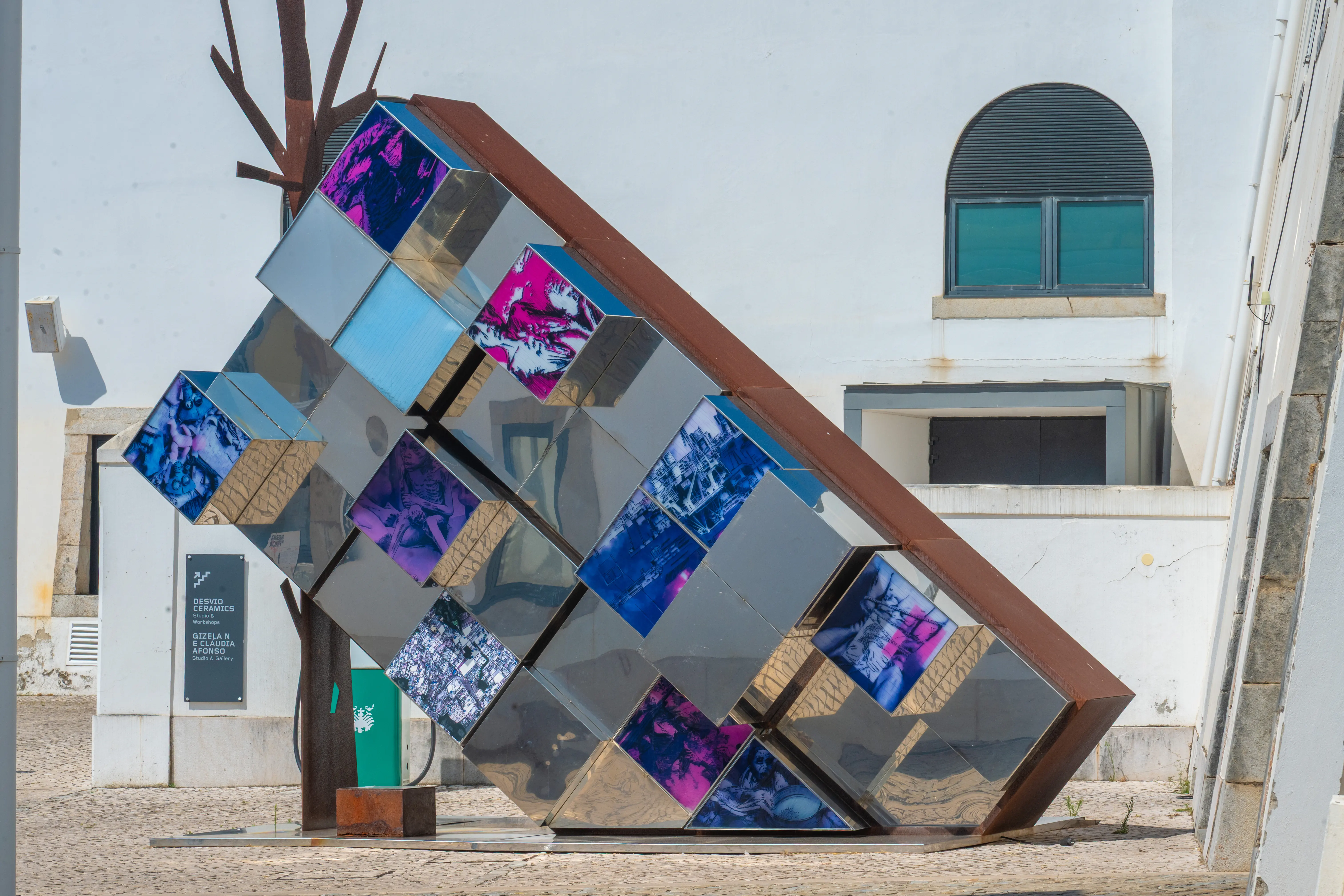 Modern outdoor sculpture made of tilted cube-like segments with colourful photographic faces, installed beside white buildings.