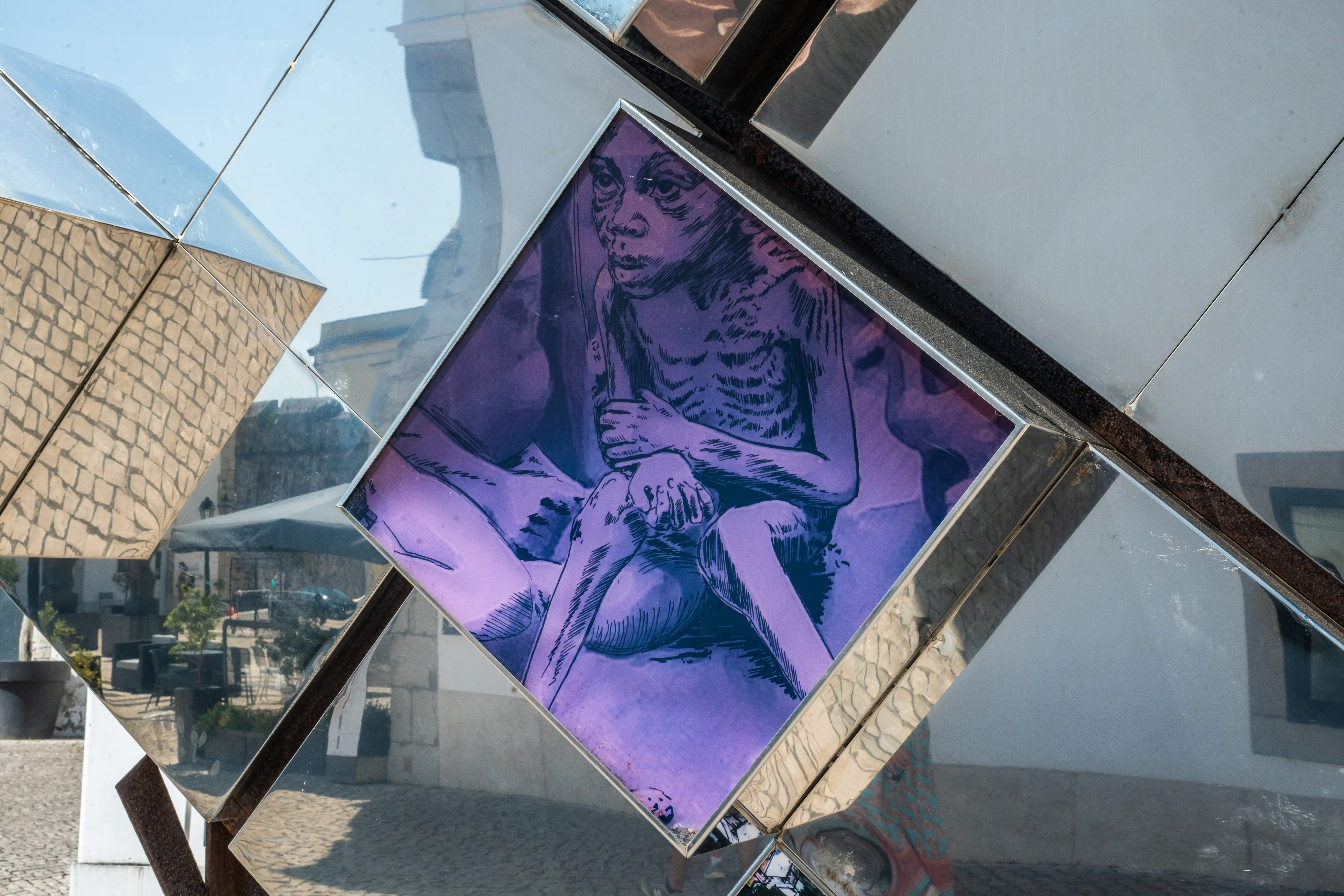 Close-up of a reflective sculpture with a central panel displaying a purple-tinted, black-and-white sketch of a skeletal figure being held. The sculpture's mirrored surfaces reflect the sky and nearby architecture.