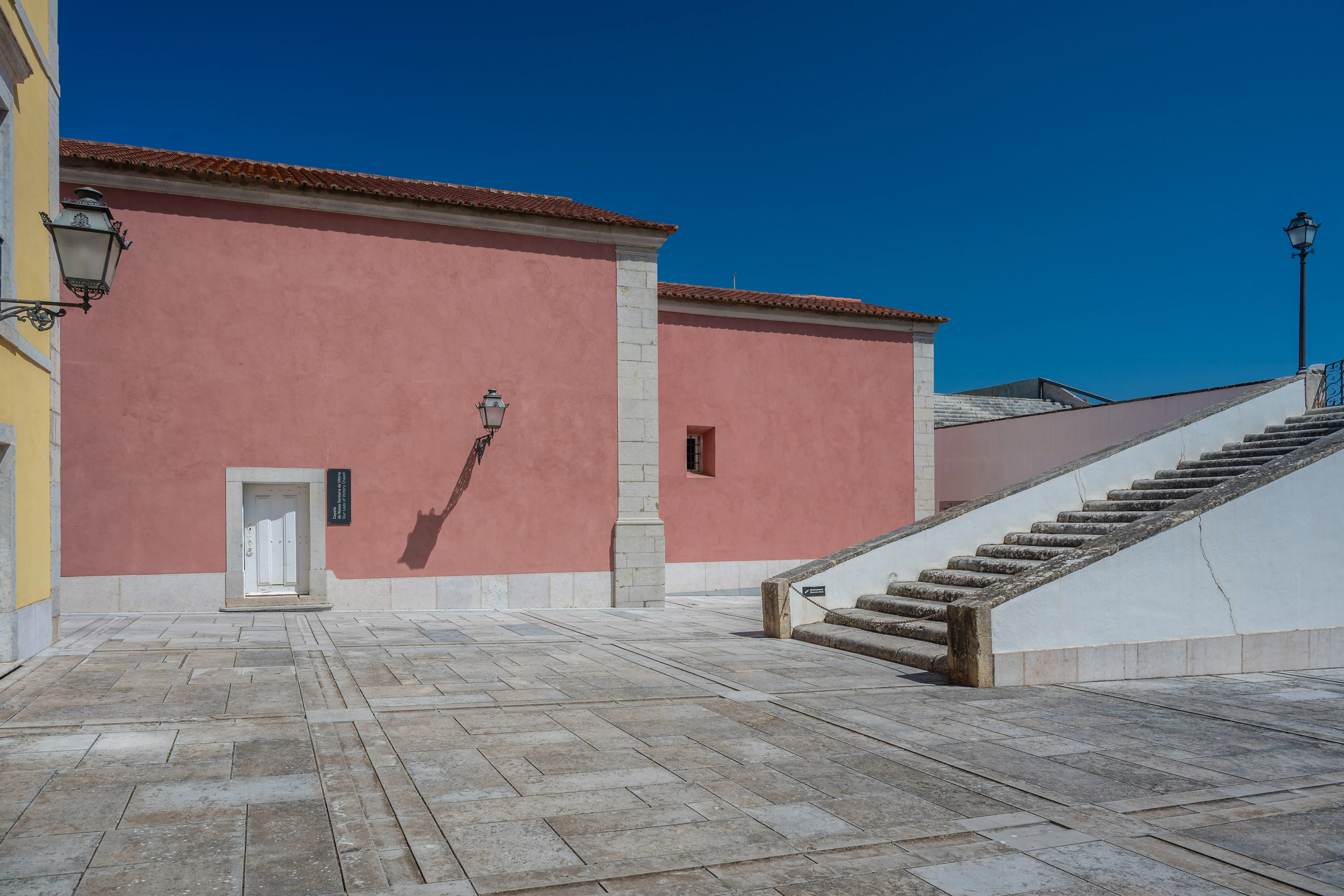 An outdoor courtyard with light grey stone paving. To the left is a salmon-pink building with a white door and a decorative black lantern. To the right, a white wall supports a wide stone staircase. The sky above is a clear, deep blue.