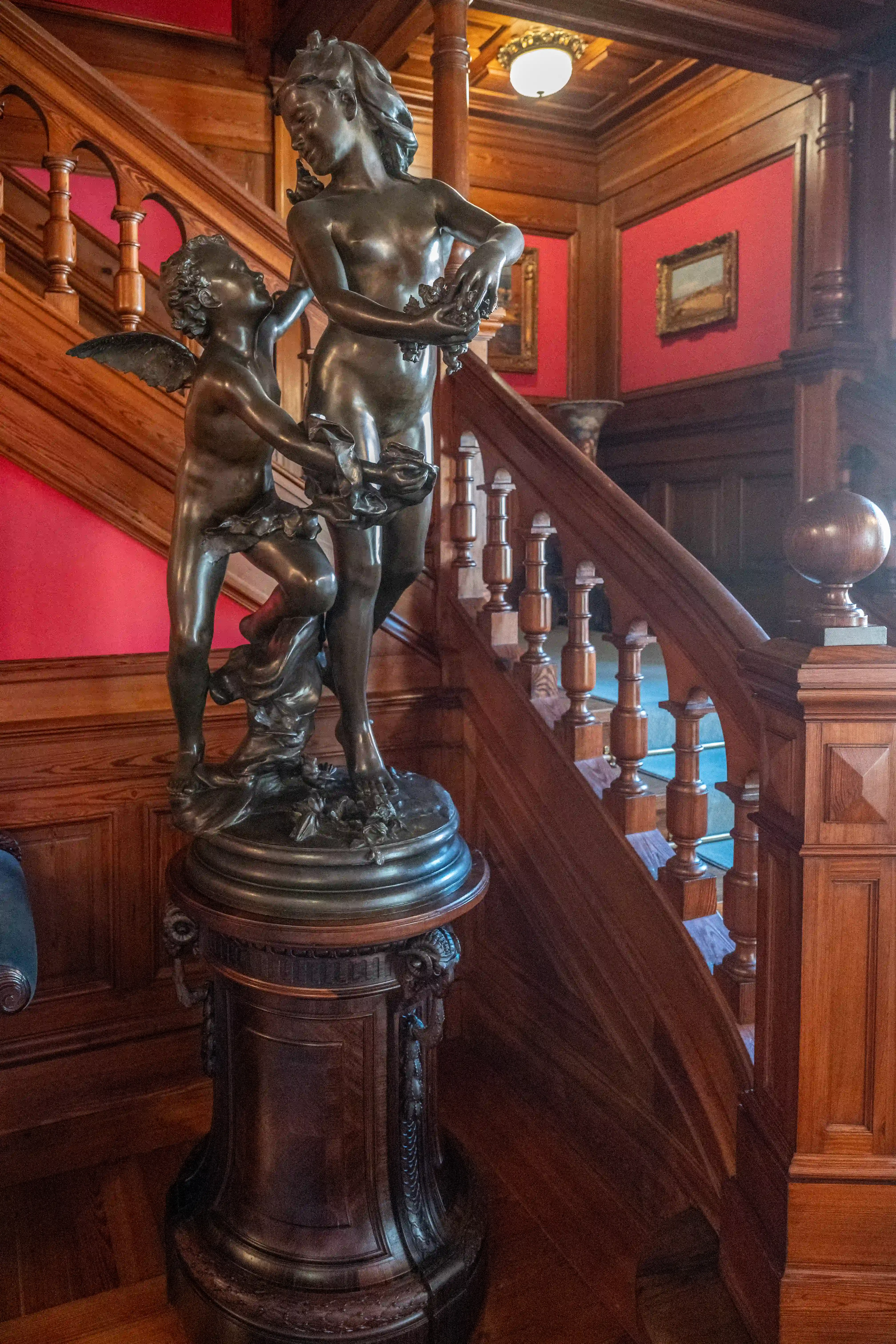 Bronze sculpture of a winged cherub and a maiden holding floral garlands, displayed on an ornate pedestal beside a polished wooden staircase with red walls.
