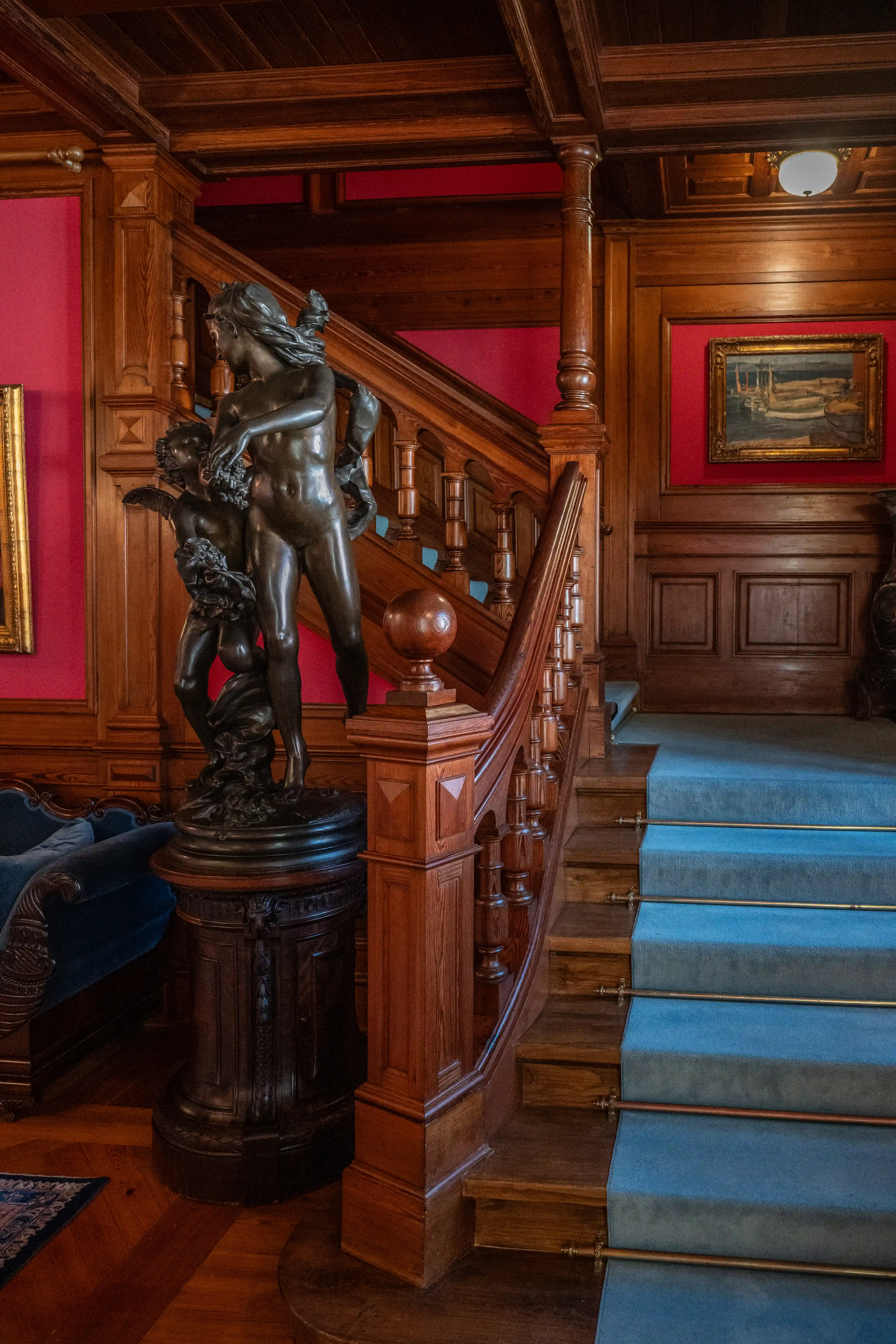 Ornate wooden staircase with blue carpet, carved banisters, and a bronze statue of intertwined figures on the landing.