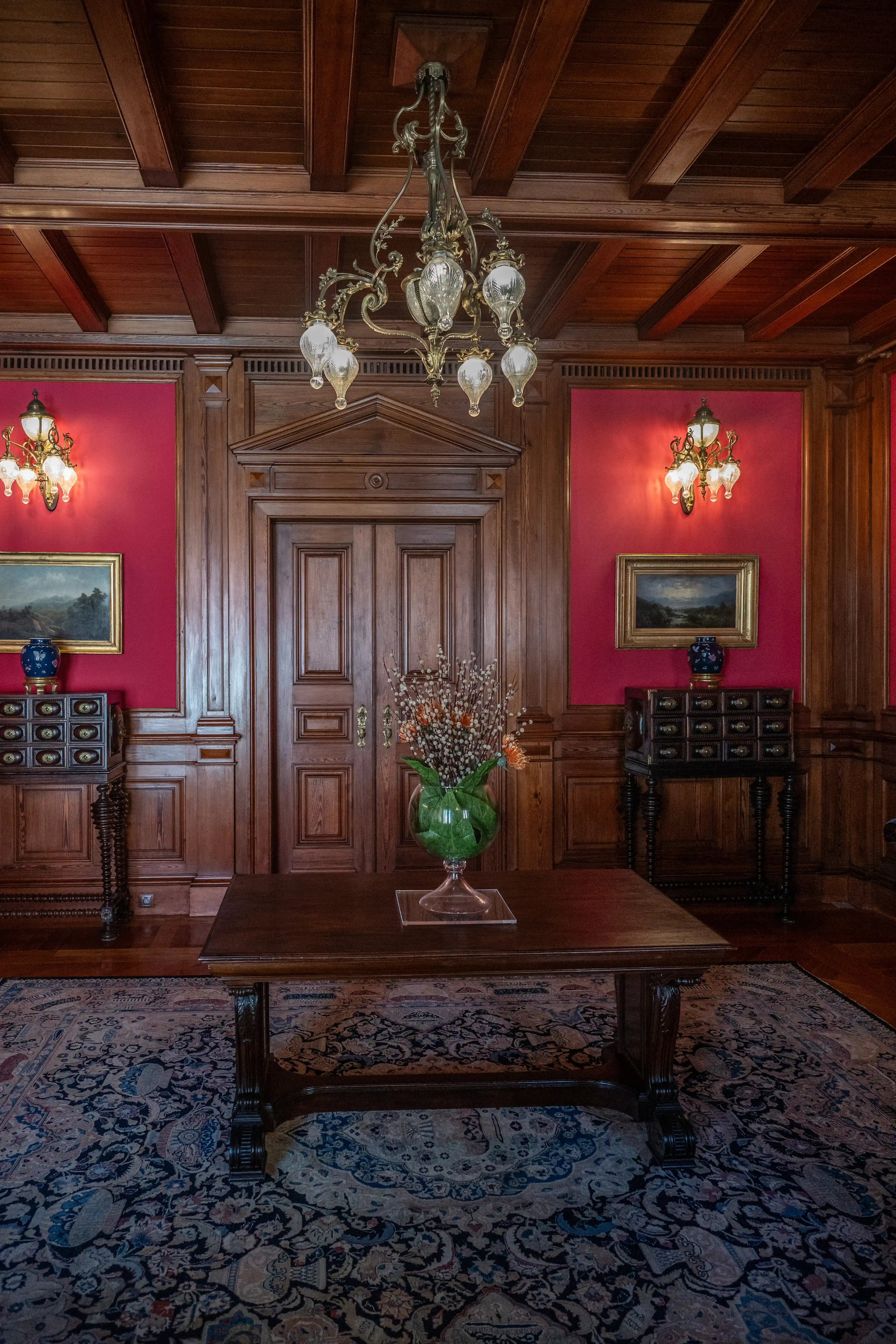 Interior view of a luxurious hallway with dark wood panelling, deep red walls, a grand chandelier, antique furniture, and a patterned rug.