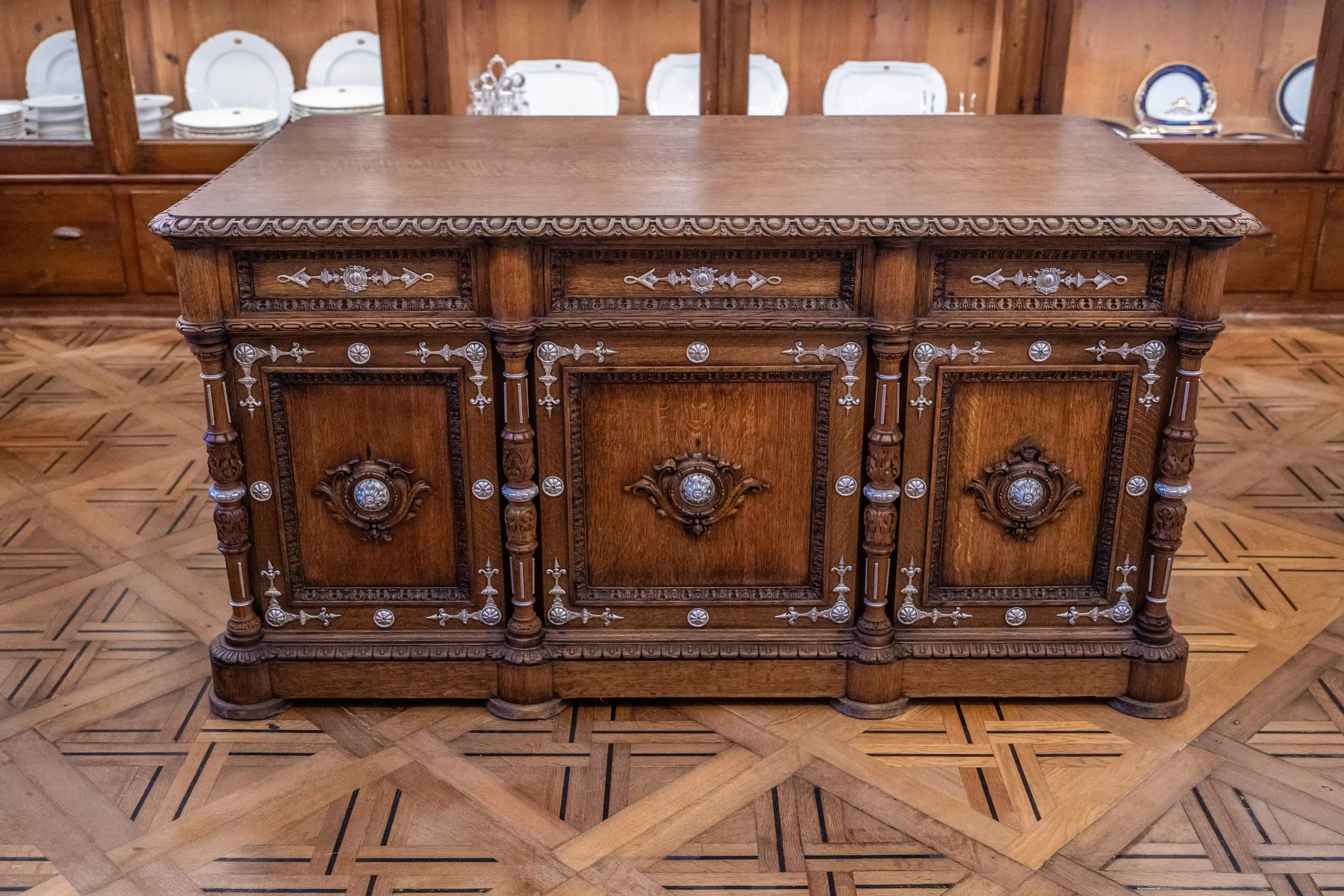 Close-up view of a large, ornate wooden sideboard with carved panels and silver decorative elements, set against a backdrop of stacked white plates in a cabinet and a patterned wooden floor.