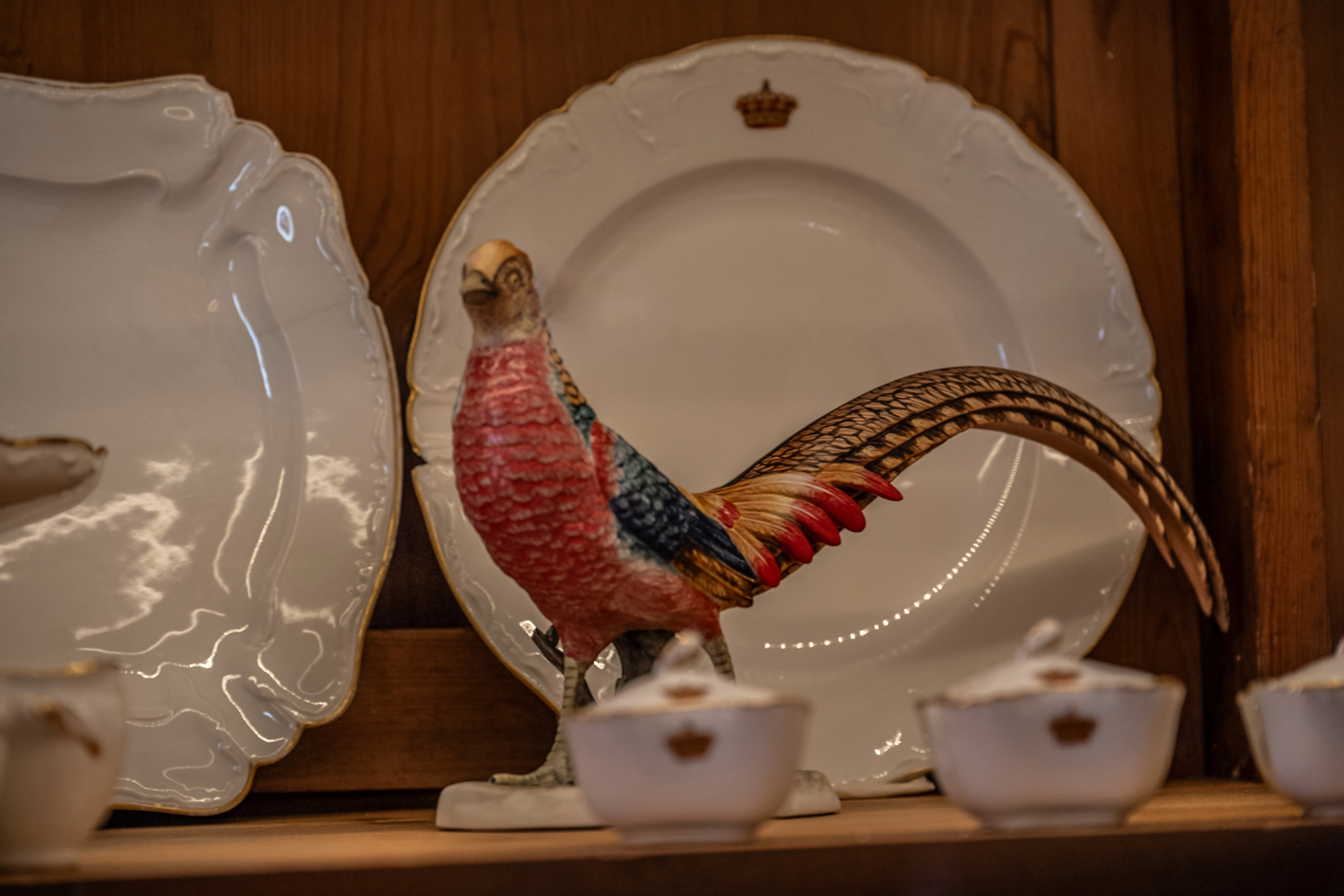 A close-up, eye-level shot of a colourful porcelain pheasant figurine positioned in front of a large decorative plate. The plate and other porcelain items in the scene feature gold trim and small crown motifs.