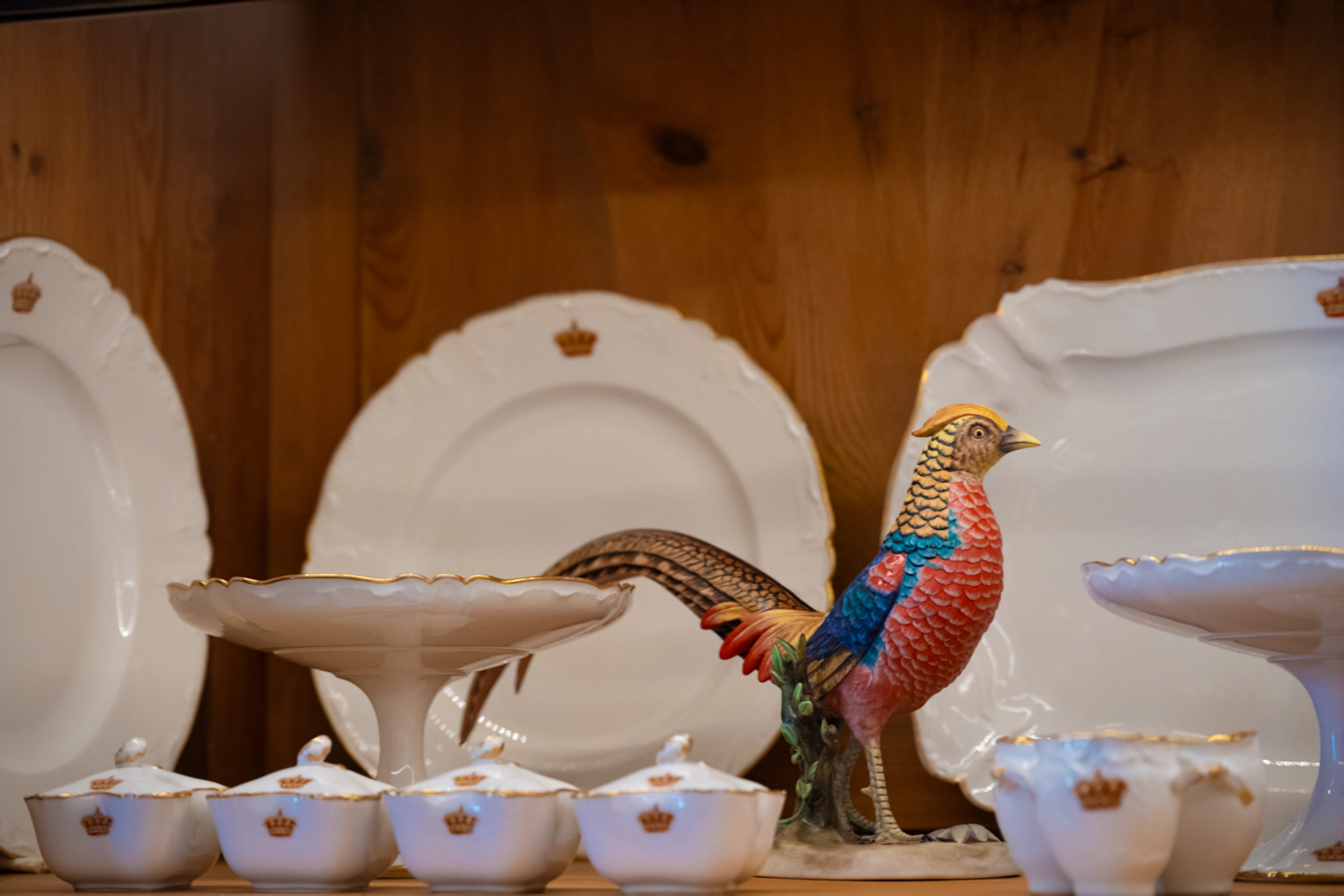 A colourful pheasant figurine is displayed in front of several white porcelain serving dishes and plates, all decorated with gold trim and a crown emblem.