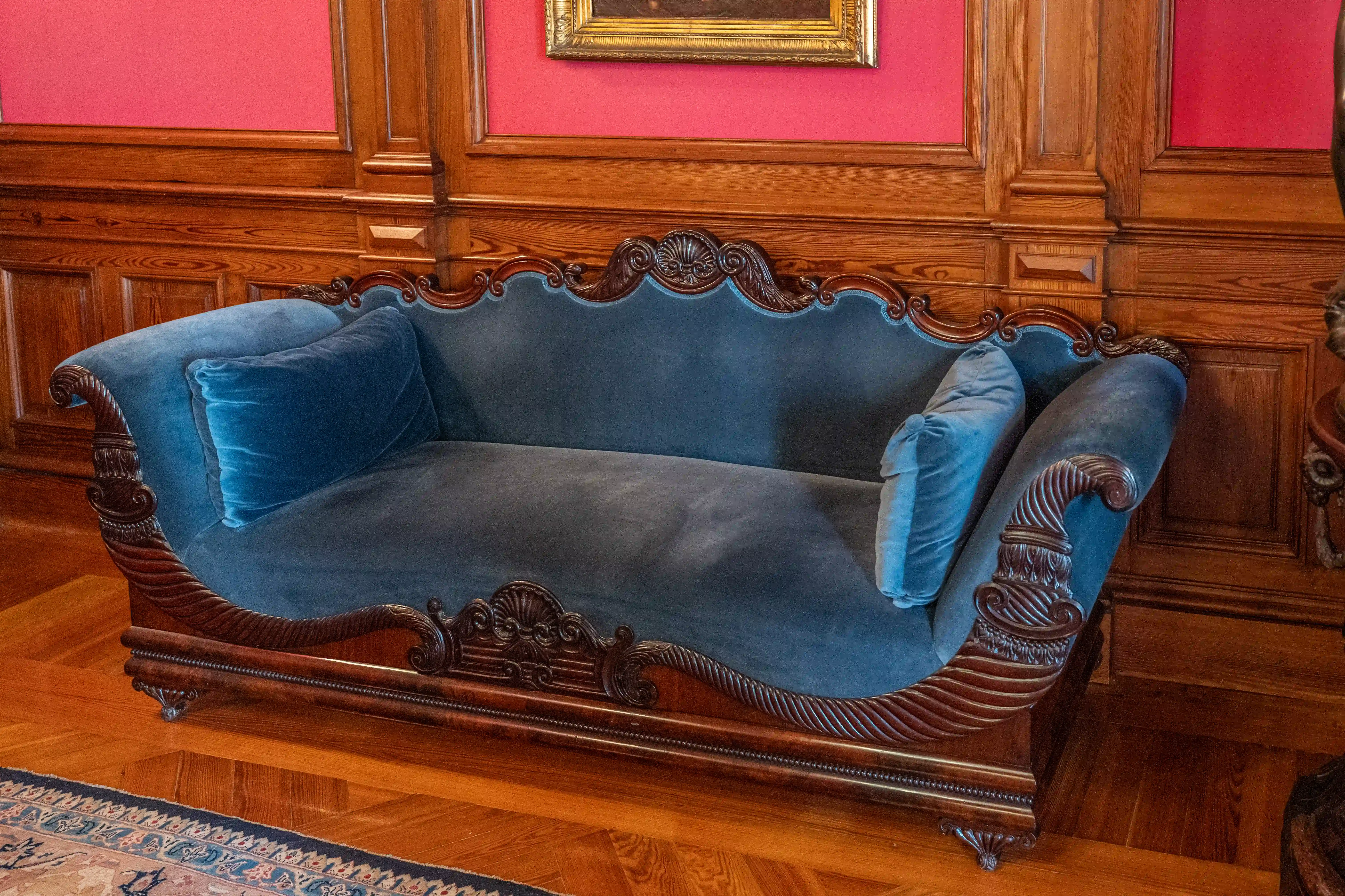 A detailed shot of a curved, dark blue velvet settee with ornate dark wood carvings and two cushions, positioned on a polished parquet floor in front of wood-panelled walls and a pink wall. A Persian rug is visible in the foreground, and a framed painting and a sculpture are partially visible in the background.