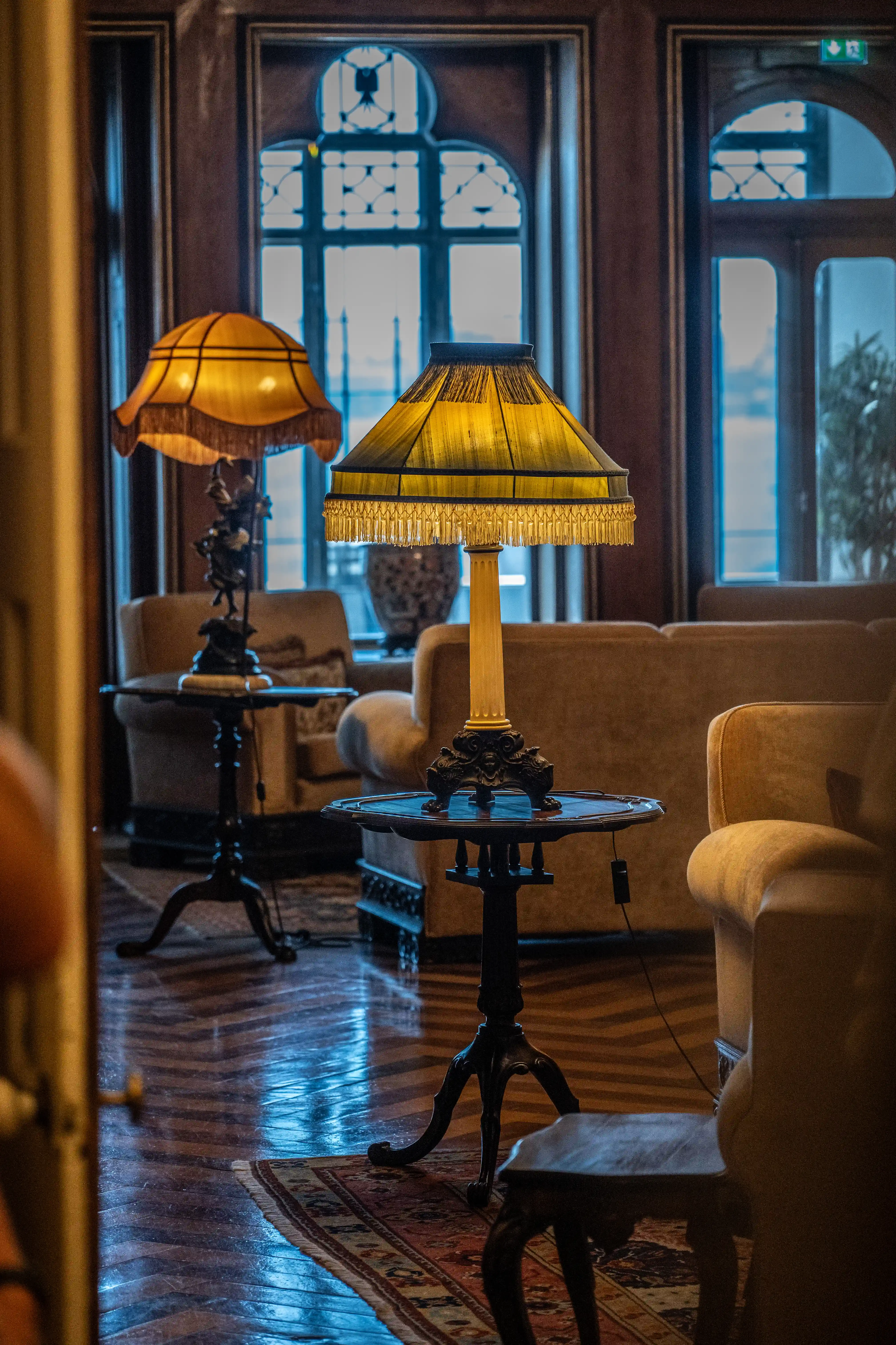 Two ornate table lamps with yellow fringed shades are positioned in a room. The lamp in the foreground is on a small table, while another is further back. Comfortable-looking sofas are visible behind the lamps, and large, arched windows with a blueish light fill the background. The floor is made of polished wood with a rug partly visible.