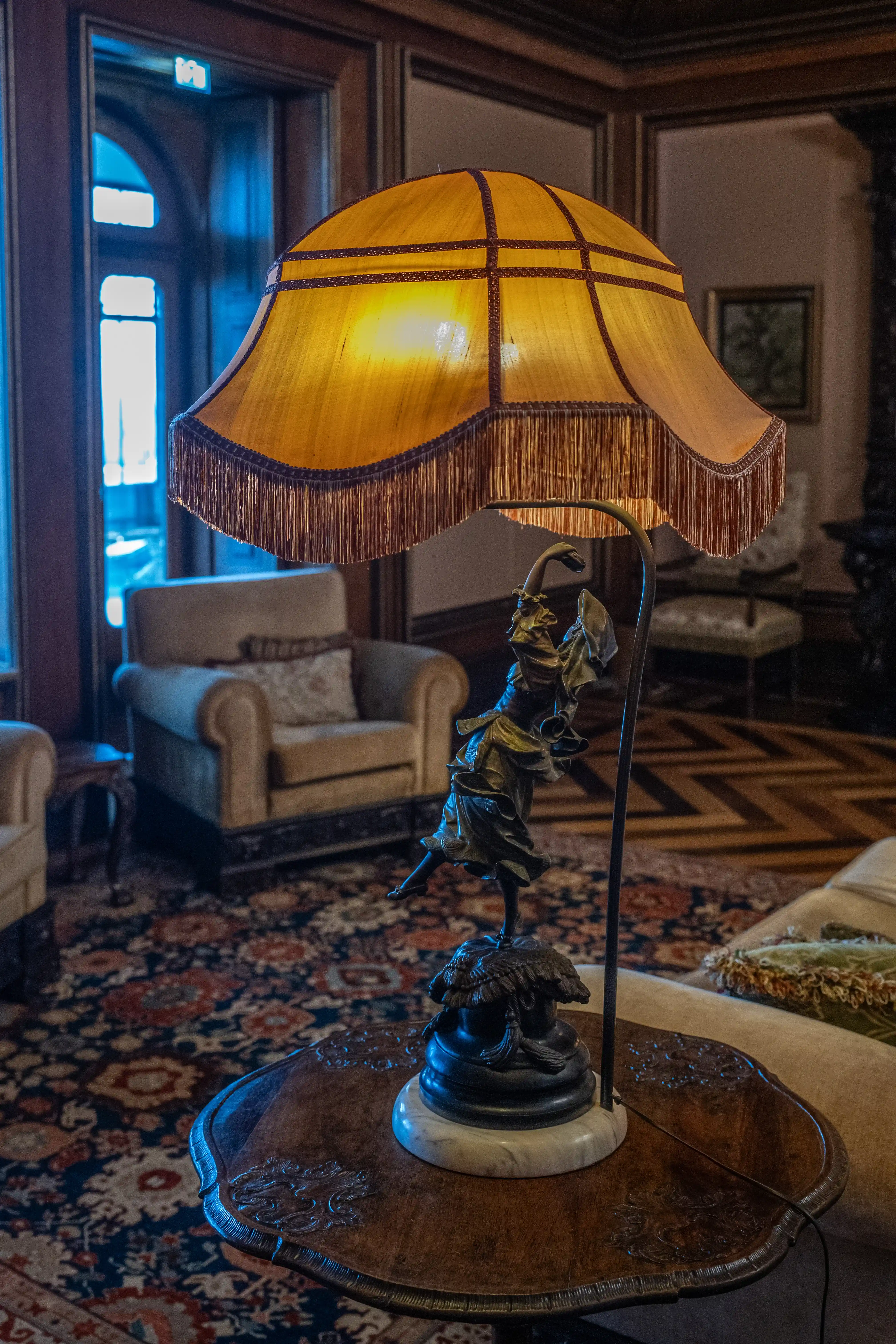 A vintage lamp with a fringed, amber lampshade and a bronze dancer figurine base on a wooden table, in a room with armchairs and a patterned rug.