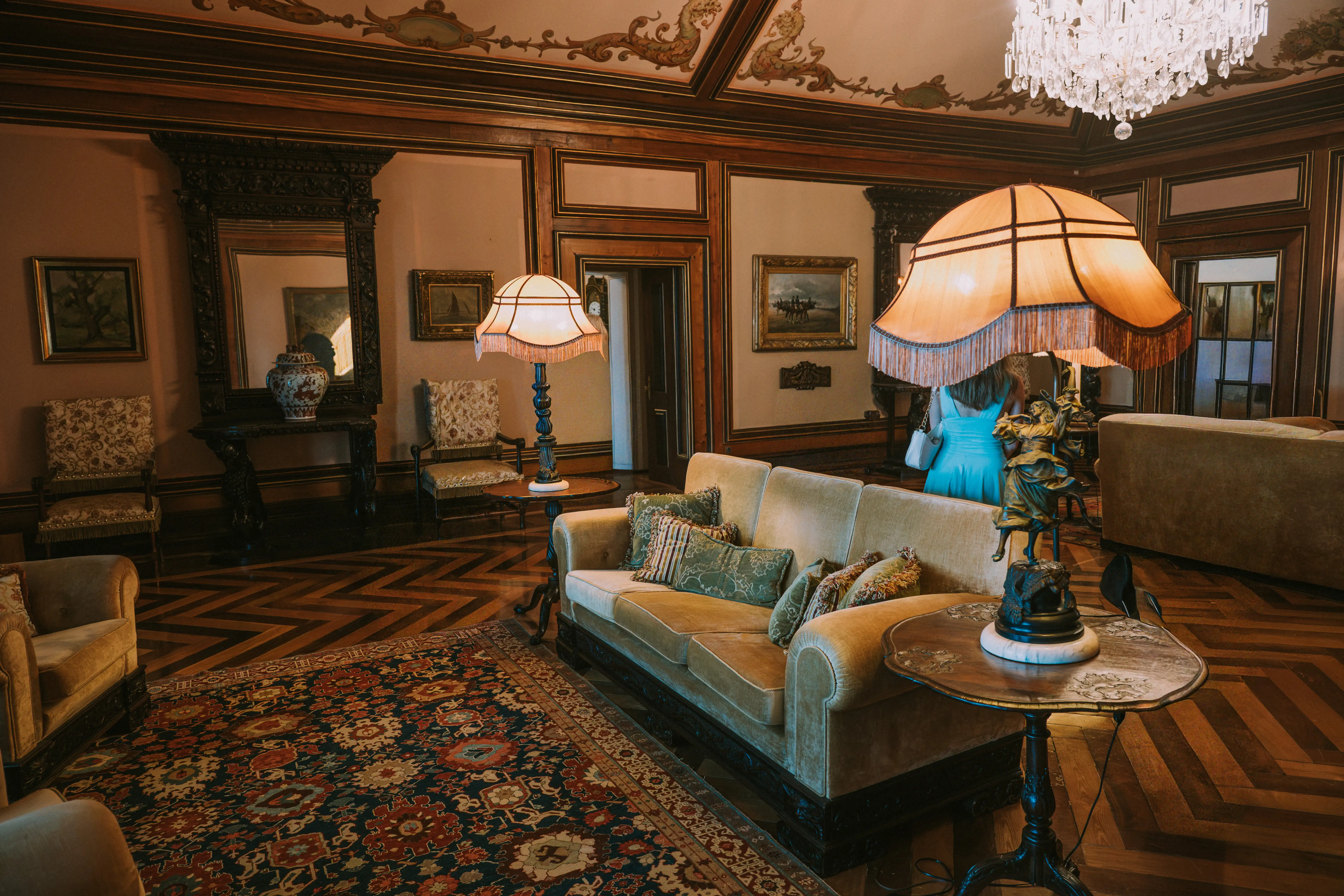 Several people are touring a well-decorated room in a historic house, examining the antique furniture and ornate lamps.