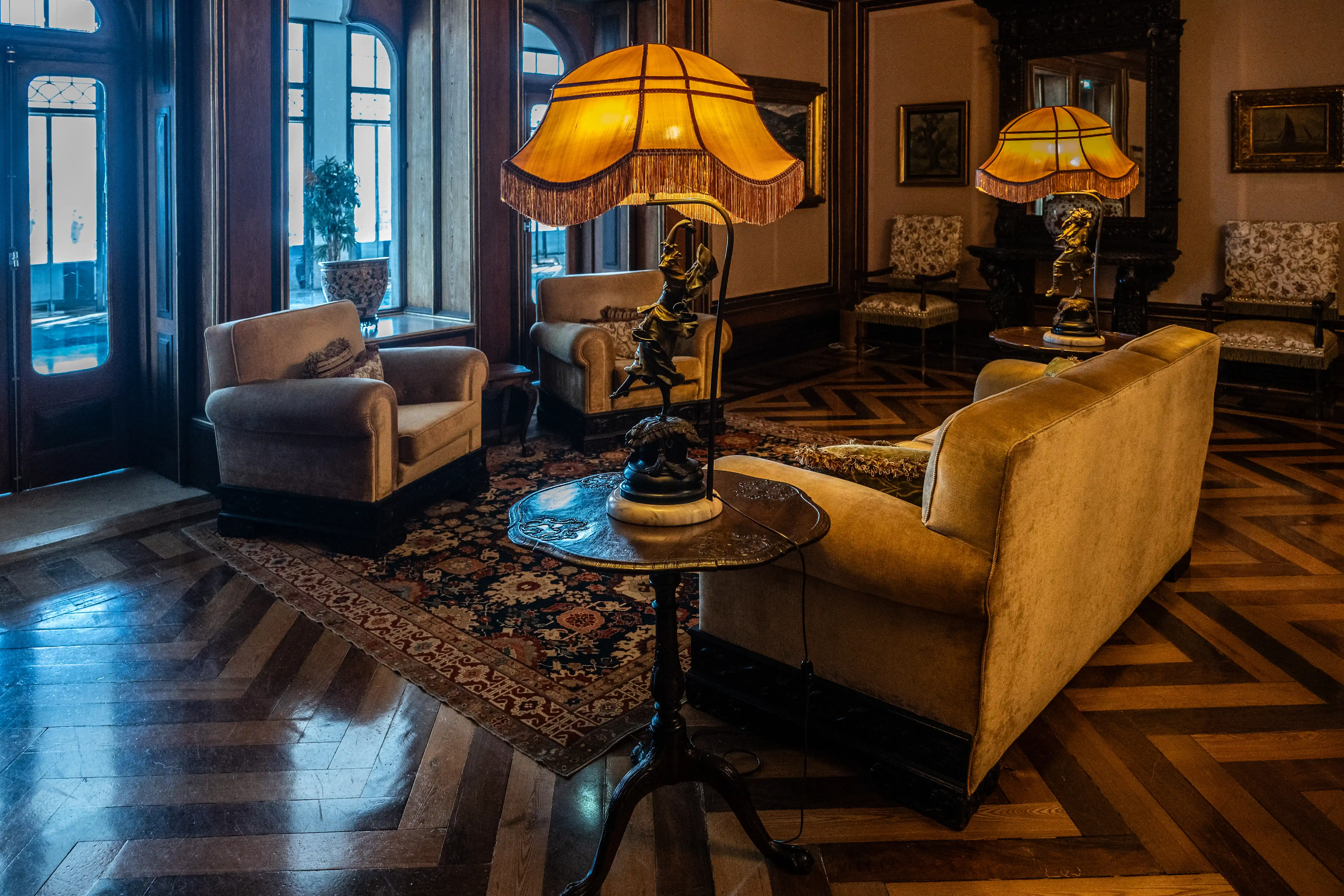 Interior view of a luxurious drawing-room with yellow upholstered furniture, antique lamps, a patterned rug, and a herringbone wooden floor.