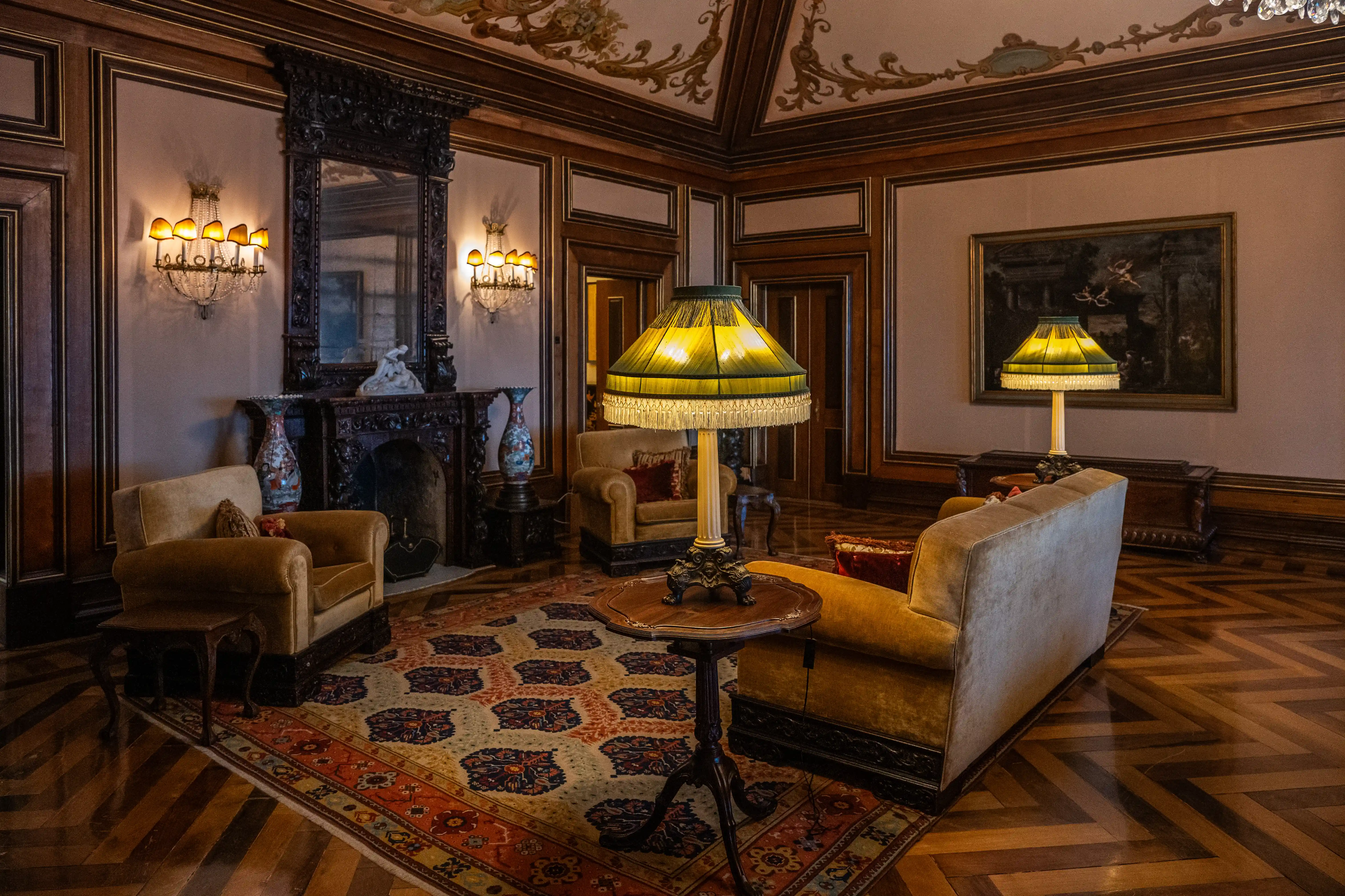 A dimly lit, opulent interior of a room with dark wood panelling, a large ornate fireplace with a mirror above, two plush armchairs and a sofa, two vintage table lamps with fringed shades, a patterned rug, and a herringbone wooden floor.