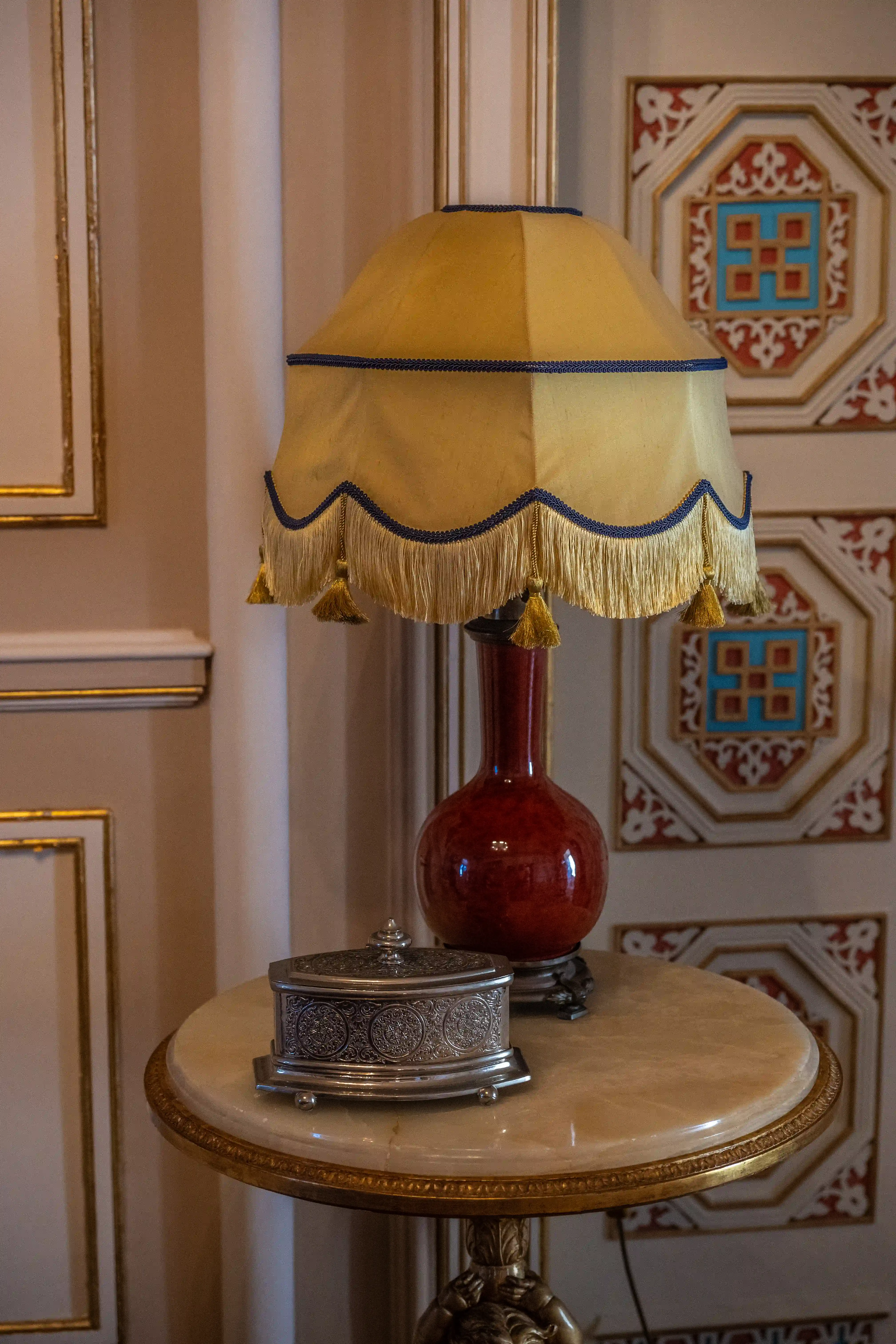 Close-up of a decorative table lamp with a gold shade and tassels, and an ornate silver box on a marble table.