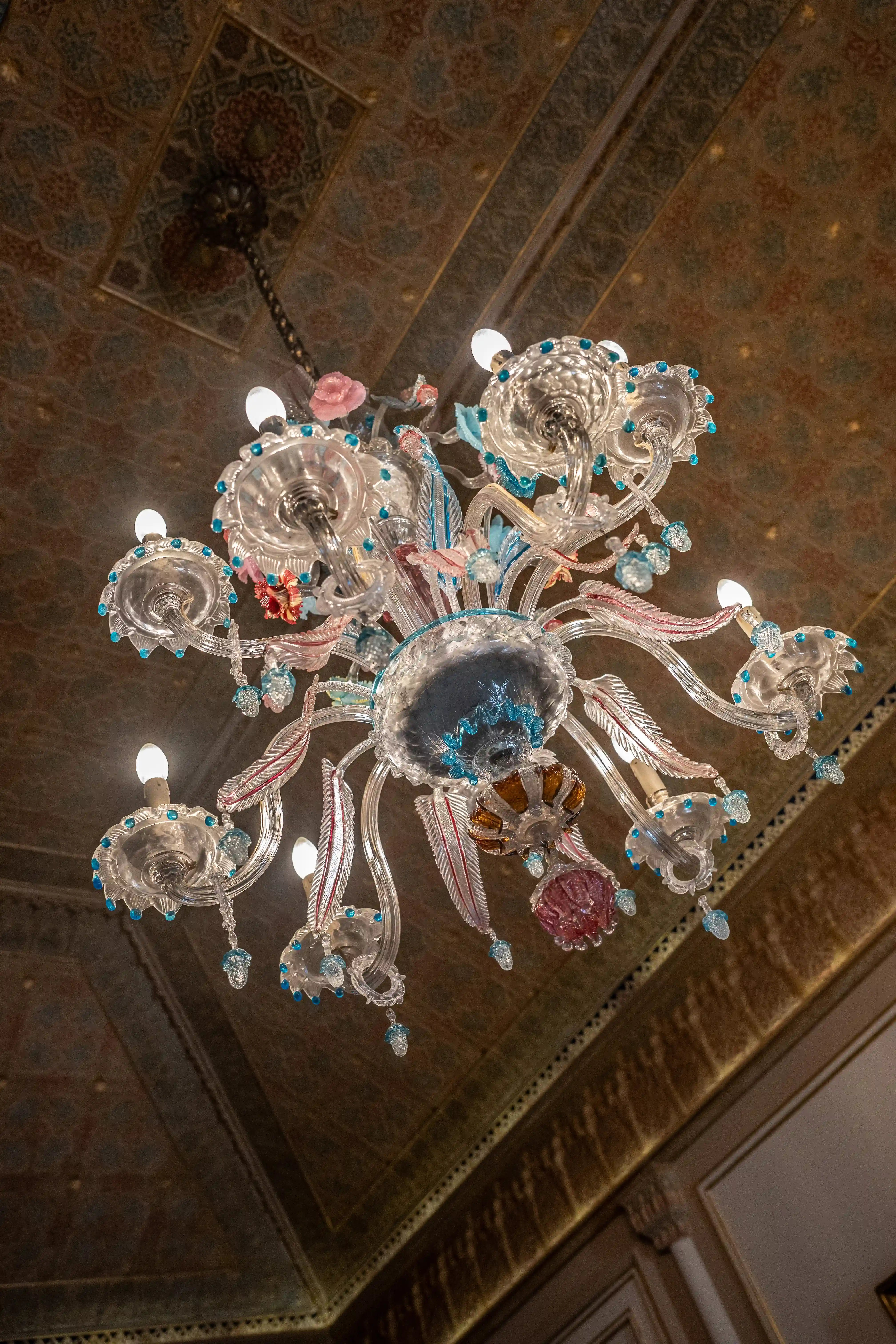 A close-up, low-angle view of a detailed Murano glass chandelier with clear, pink, and blue glass elements, hanging from a richly patterned ceiling.
