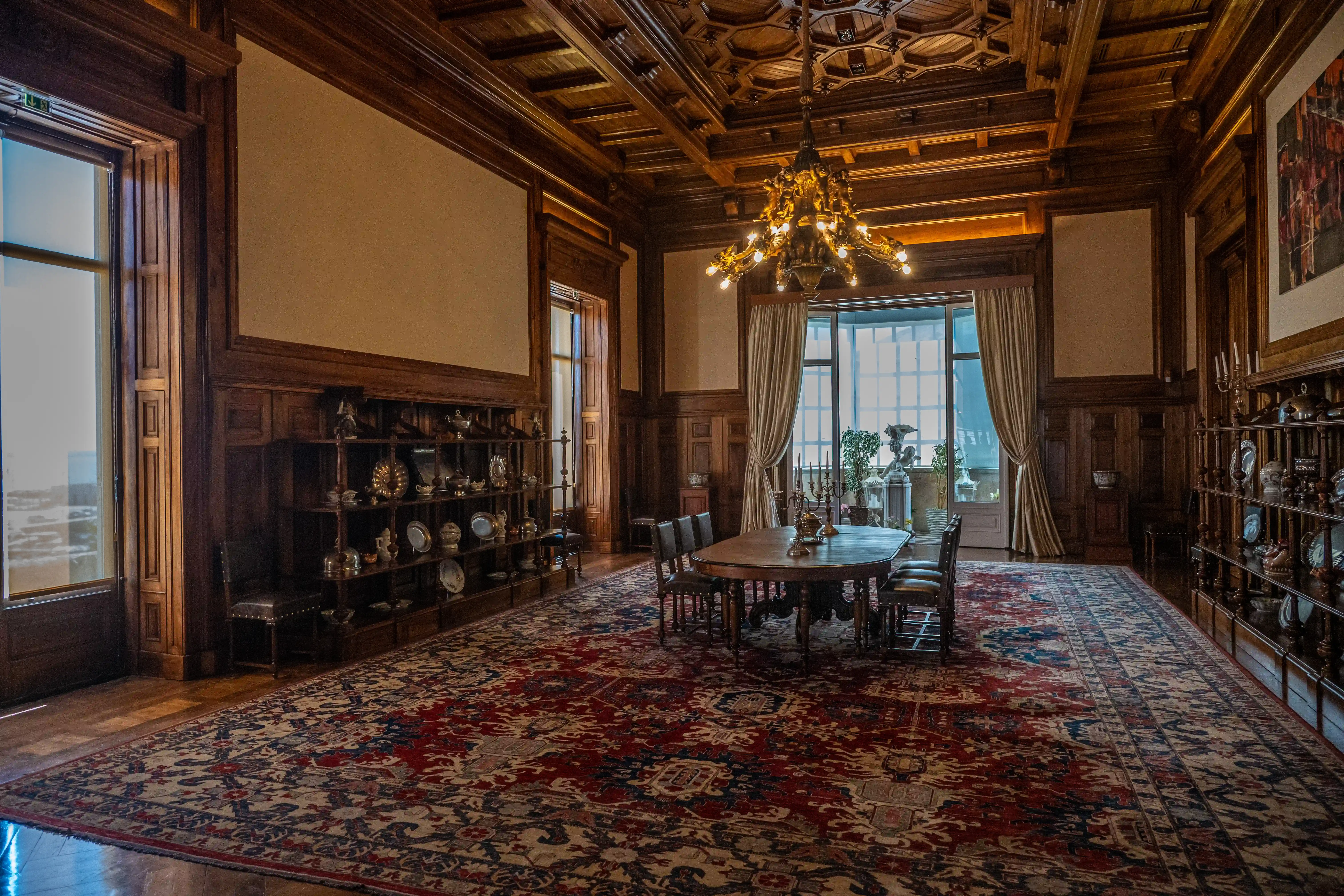 Interior view of an opulent dining room with dark wood panelling, an ornate ceiling, a large patterned rug, a dining table set for six, and shelves displaying antique tableware.