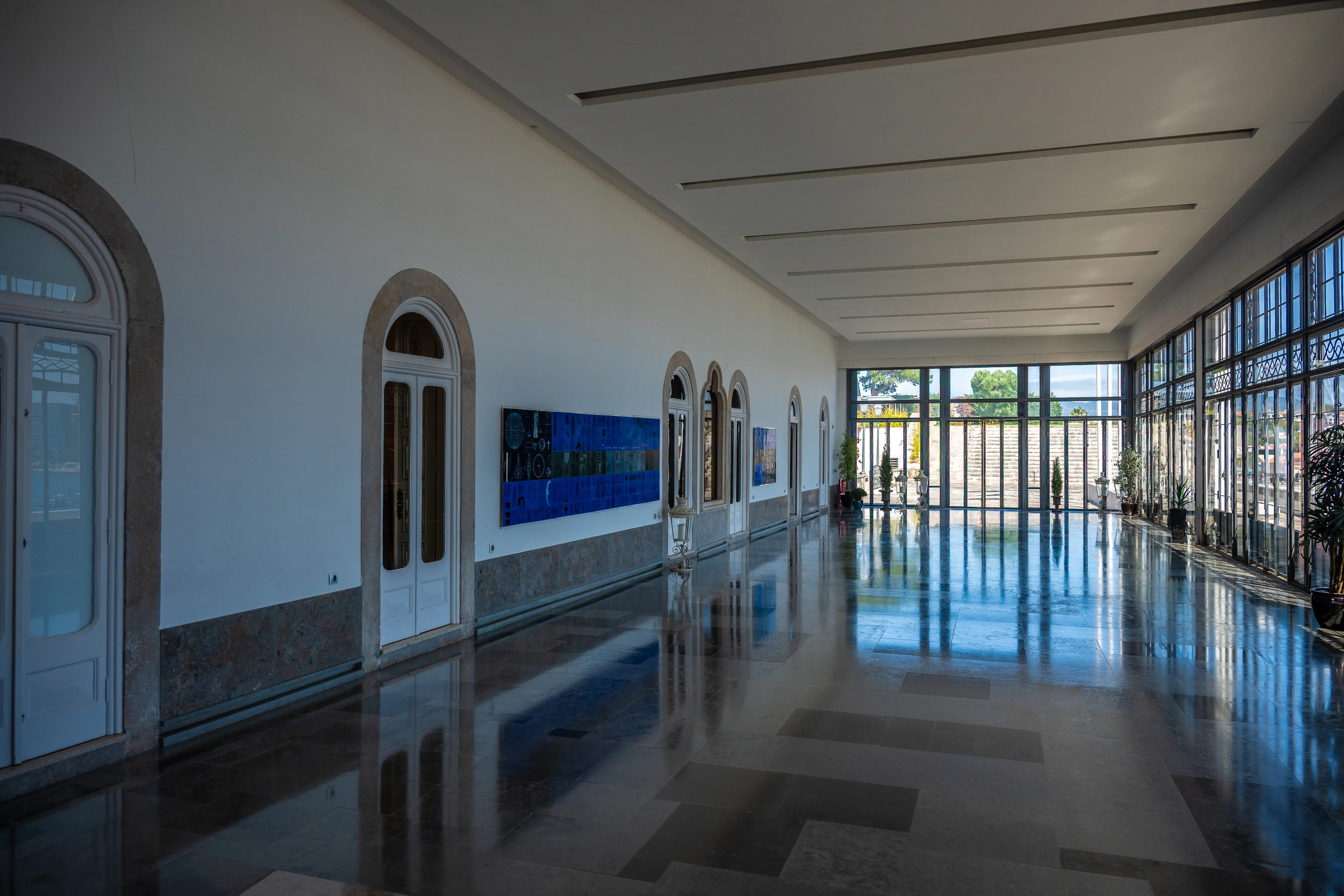 A spacious hall with a polished marble floor reflecting light from large arched windows and glass doors. The walls are white with a gray marble base, and arched doorways and modern art pieces adorn the space