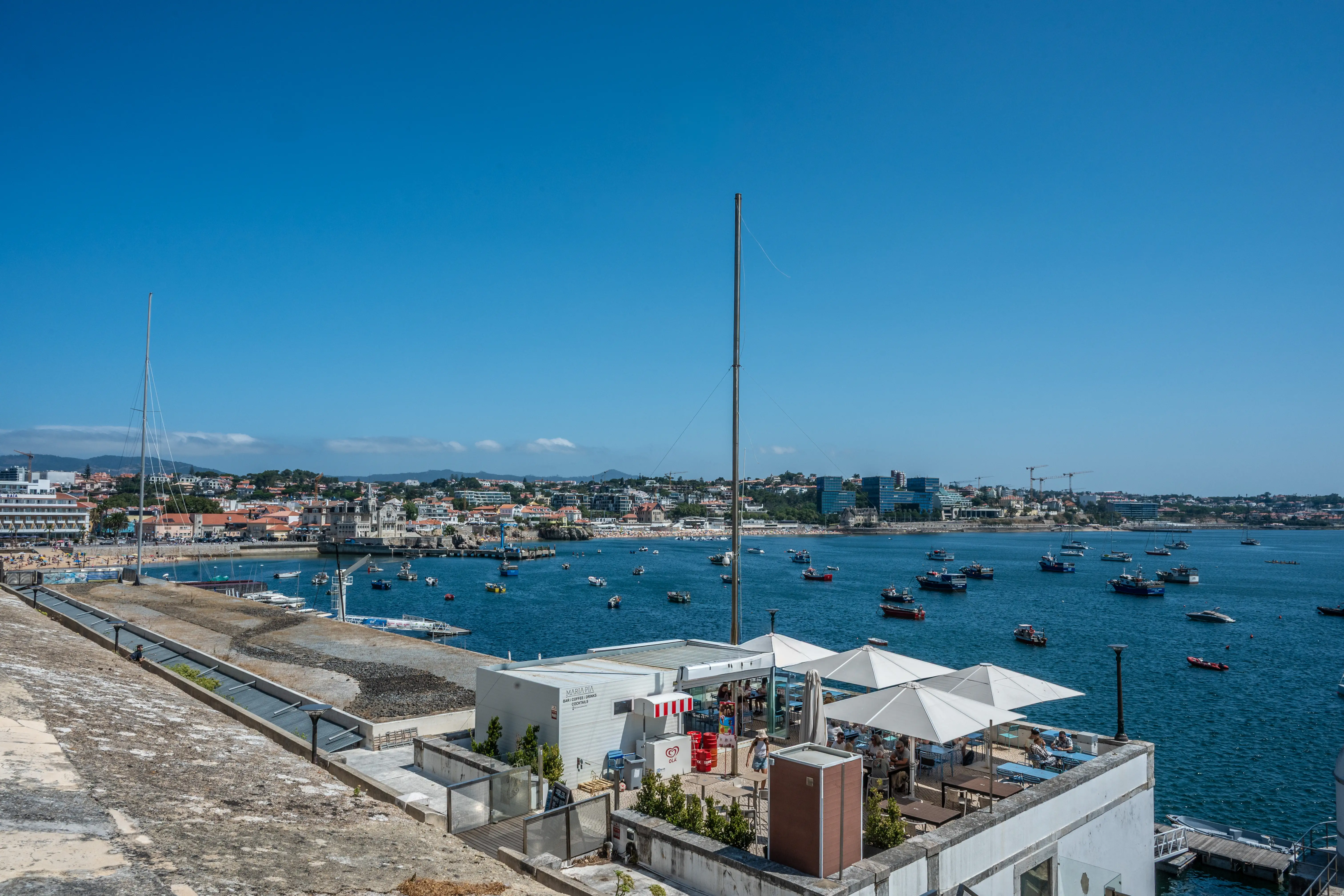 A wide shot of a bay filled with boats, with a town and buildings in the background. In the foreground, there is an outdoor cafe with white umbrellas and seating.