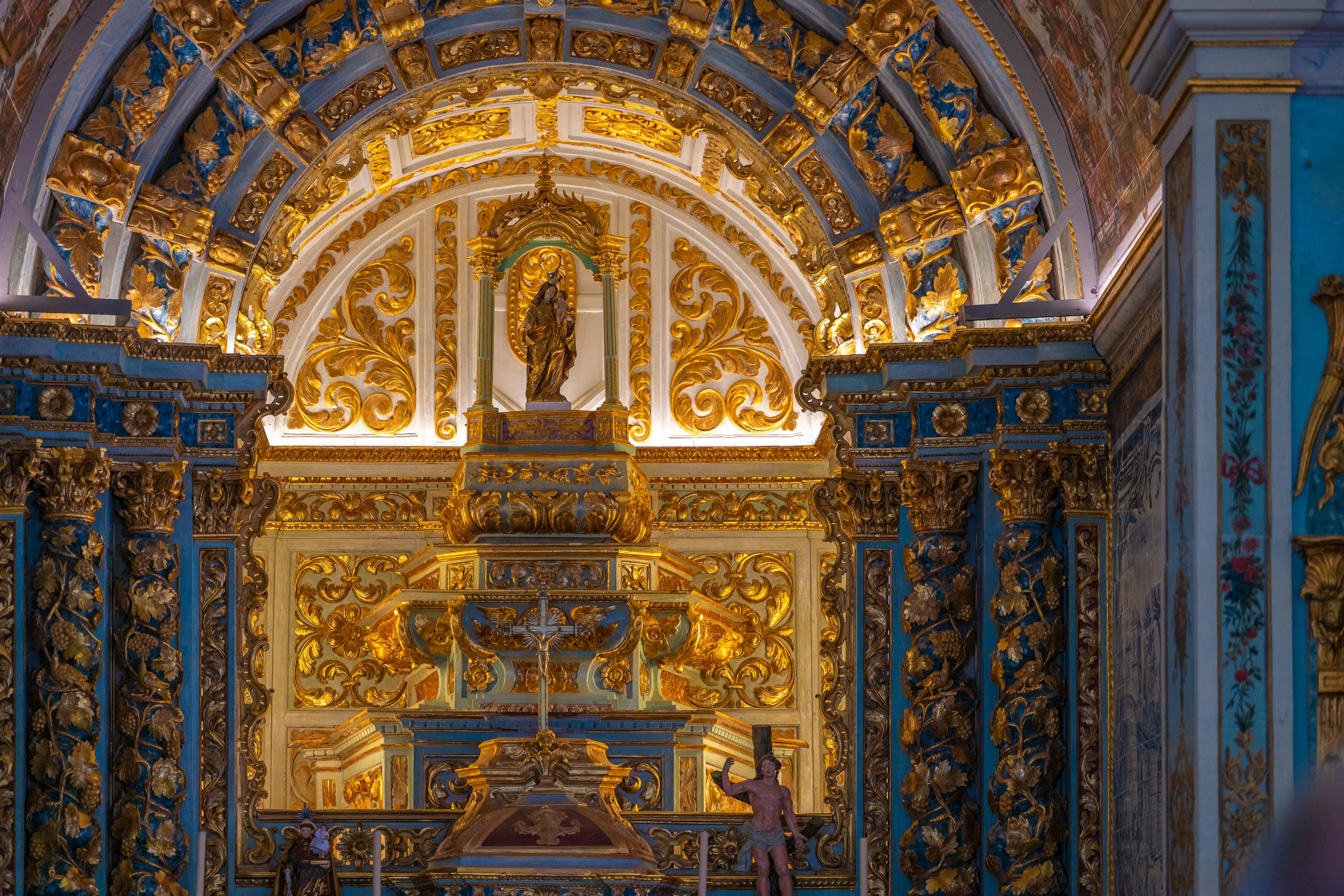 Interior view of a church altar, with elaborate gold-gilded carvings, blue painted details, and religious statues, characteristic of Baroque architecture.