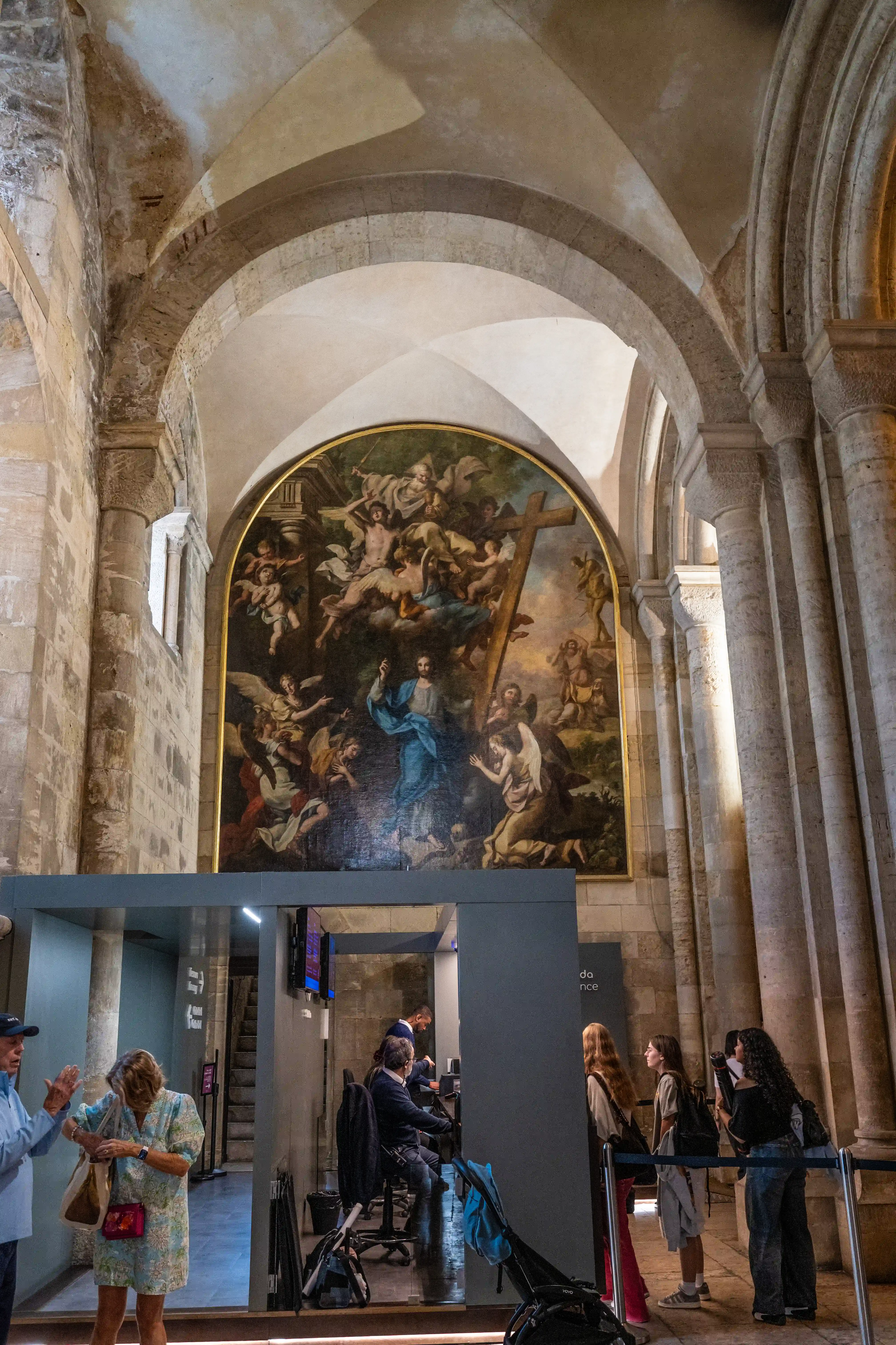  Interior view of a grand cathedral with a large, ornate religious fresco on the far wall. In the foreground, there is a modern exhibition booth with several people.