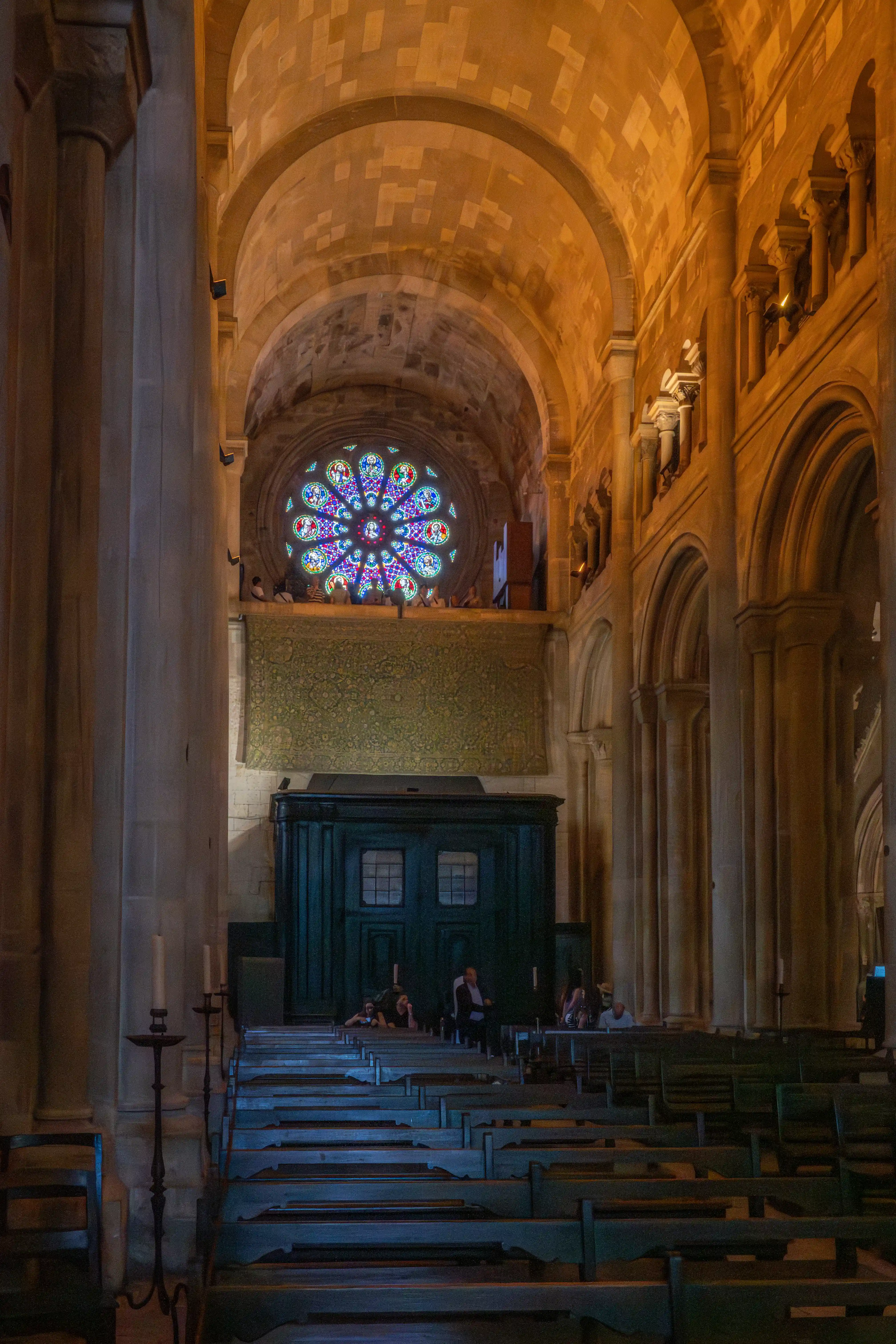  Interior of a grand cathedral featuring a colourful stained-glass rose window above a dark green wooden structure, with rows of pews and large stone columns