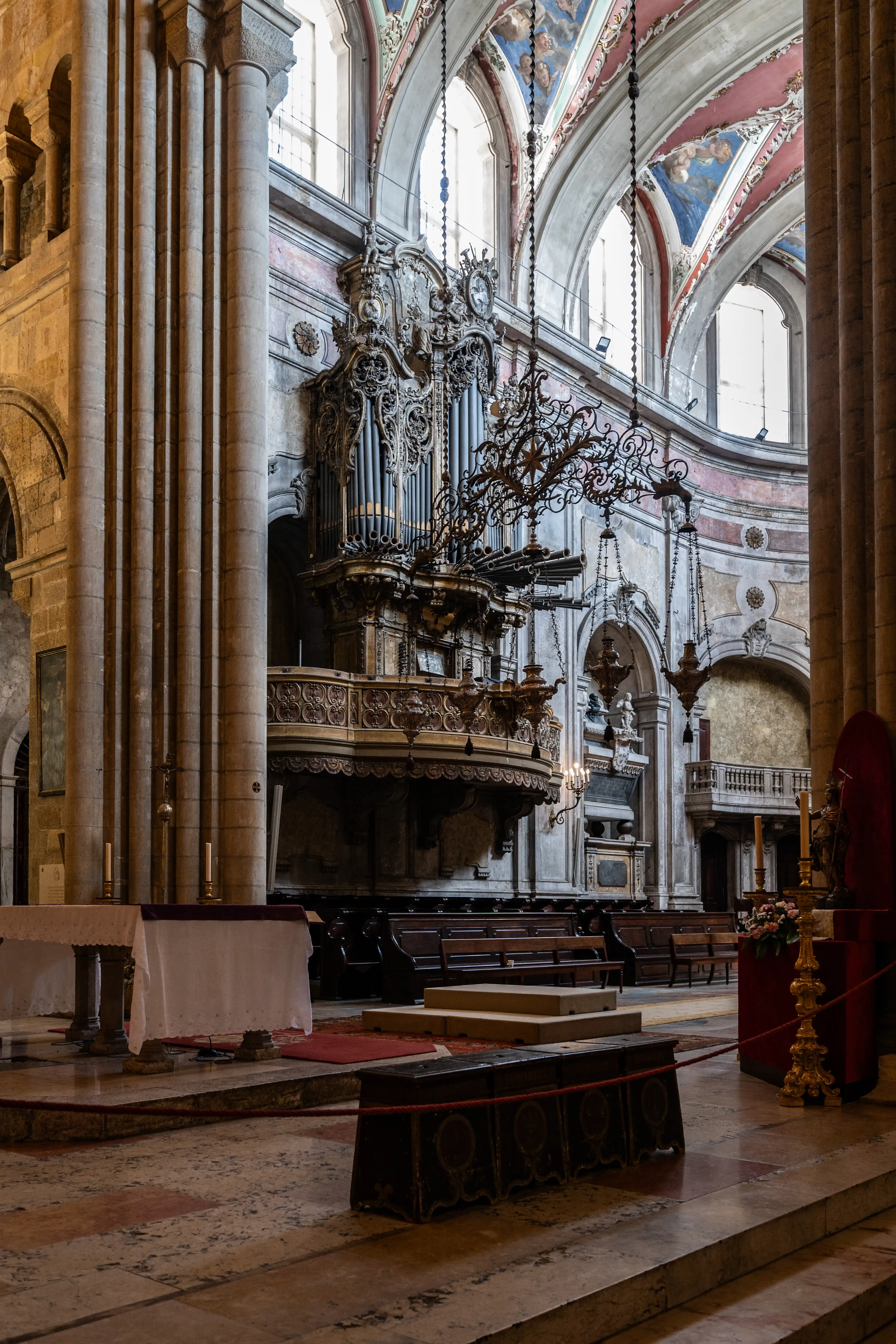 A wide-angle shot captures the ornate interior of a cathedral, dominated by a magnificent pipe organ mounted on a balcony. The organ's intricate golden detailing and rows of pipes are illuminated by natural light streaming through arched windows. In the foreground, an altar draped with a white cloth and adorned with candles sits on a raised platform. The scene also features carved wooden pews, decorative chandeliers, and frescoes adorning the vaulted ceiling, showcasing the grandeur and historical significance of the religious edifice.
