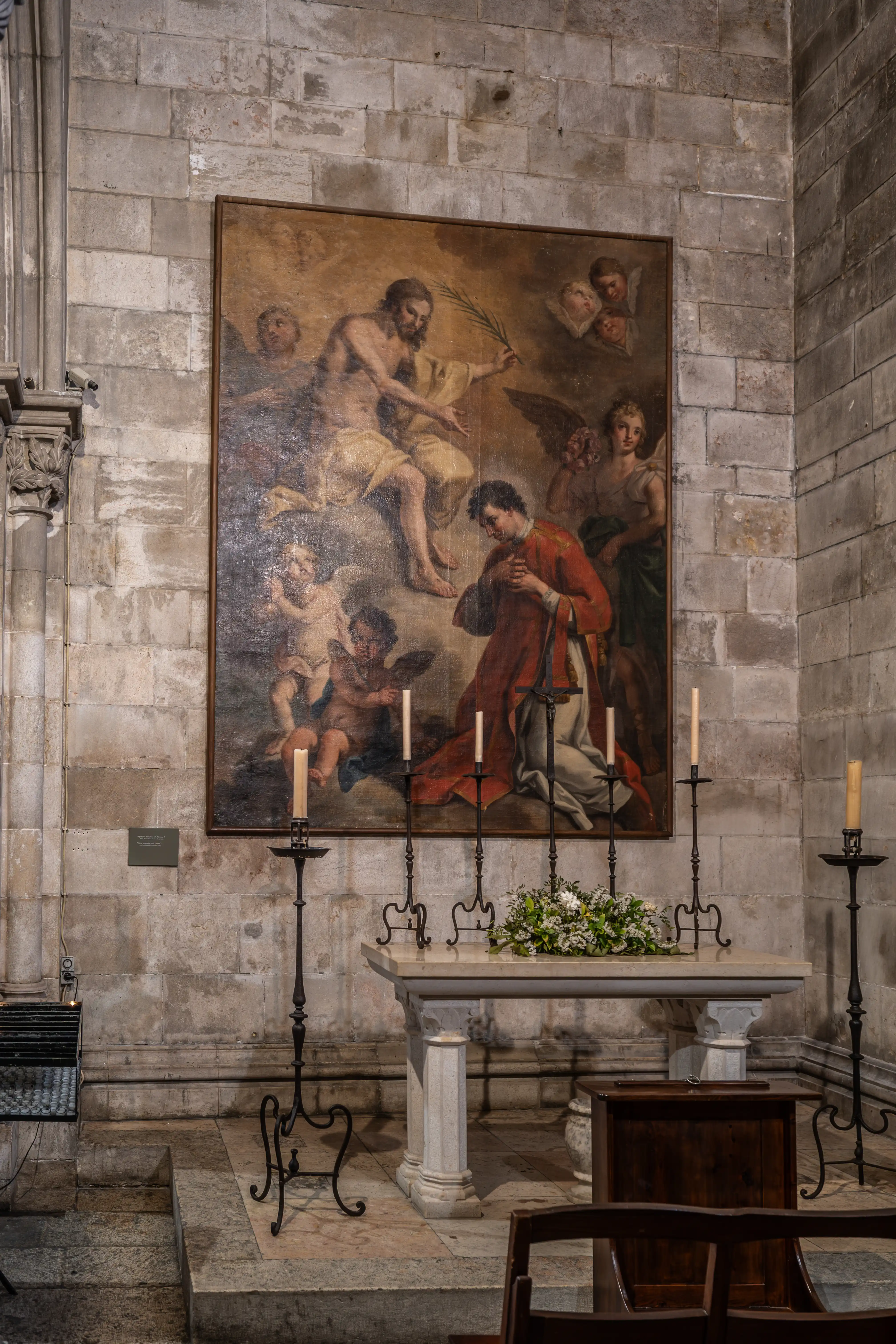 An ornate oil painting in a church depicts a bearded man in a toga offering a palm frond to a kneeling man in a red robe, with cherubs and an angel above. An altar with flowers and lit candles stands before the painting in the stone-lined interior.